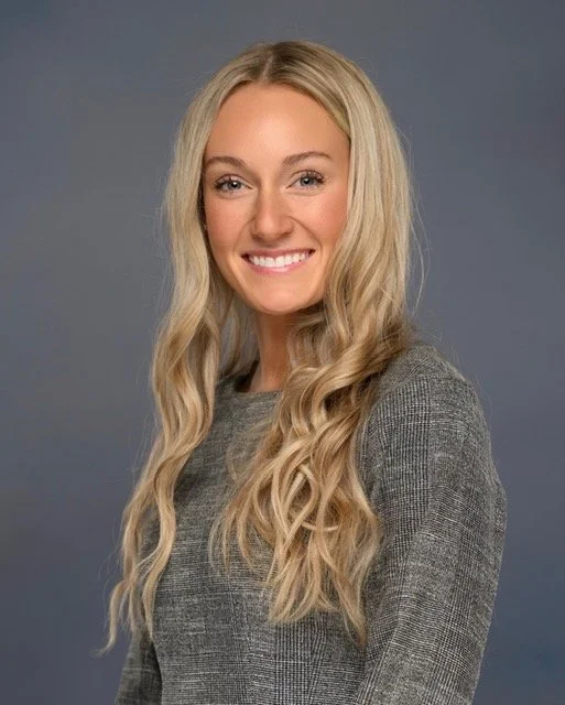 A woman sitting and smiling, she has long blonde hair, lightly tanned skin, and is wearing a grey sweater.