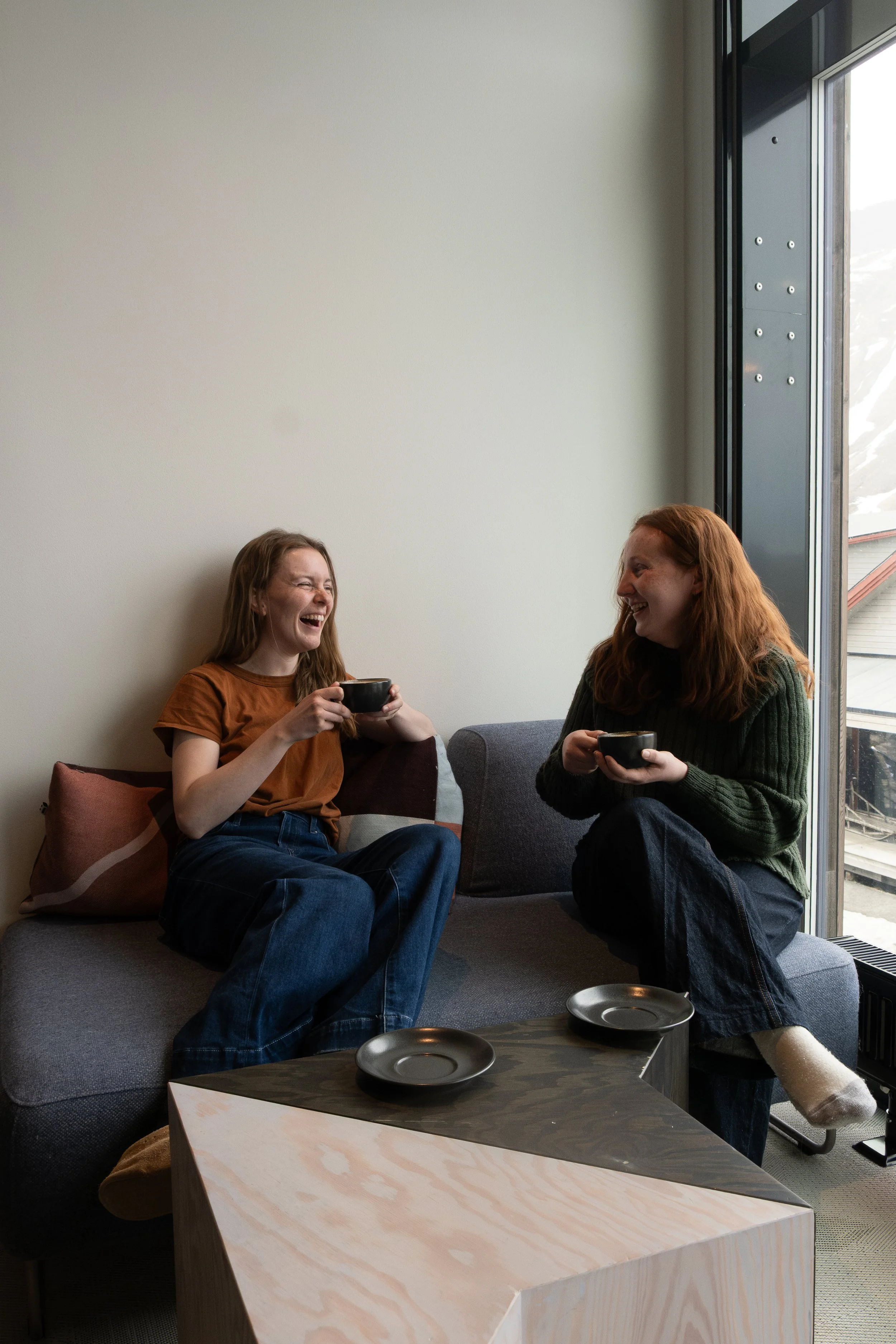 To kvinner som sitter på en sofa i et rom. Den ene kvinnen har på seg en brun t-skjorte og den andre har en grønn genser. Begge ler og holder kopper med drikke. Det er store vinduer bak dem, og en kaffebord foran.