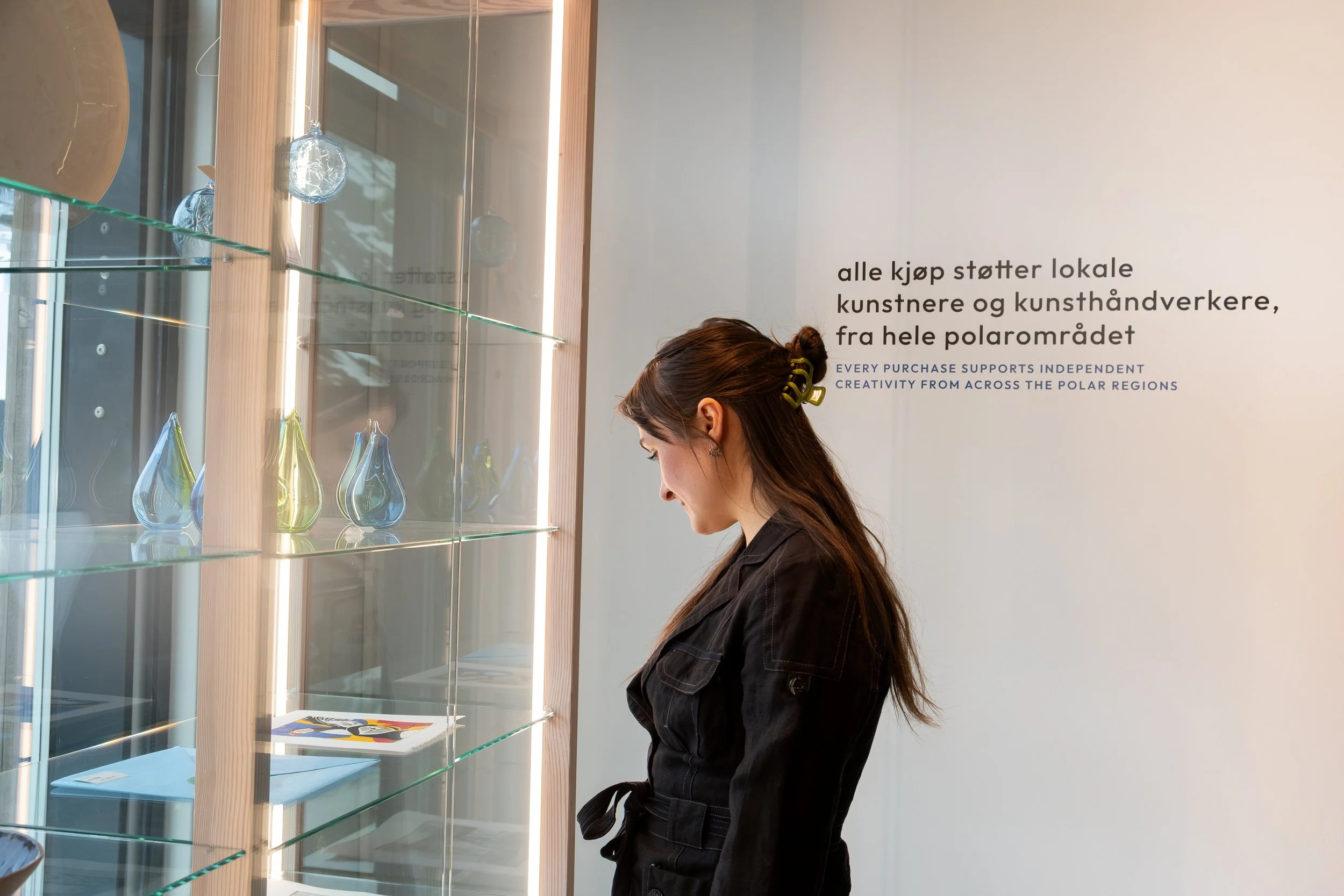 En kvinne med langt brunt hår i hestehale ser på glasskulpturer i en utstilling, med en vegg med tekst på norsk og engelsk i bakgrunnen.