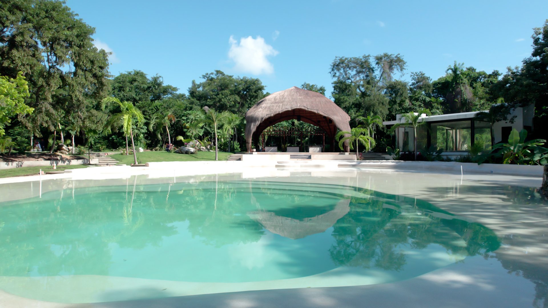Luxury pool with clear water surrounded by lush greenery and tropical trees, featuring a thatched-roof hut and a modern glass structure in the background, under a bright blue sky.