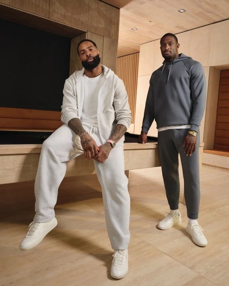 Two men dressed in casual athletic clothing posing in a modern room with wooden walls and floor. One man is sitting on a wooden edge, wearing a white hoodie and sweatpants, with tattoos visible and a beard. The other man is standing, wearing a gray hoodie and joggers, with a gold watch and chain. Both are wearing white sneakers.