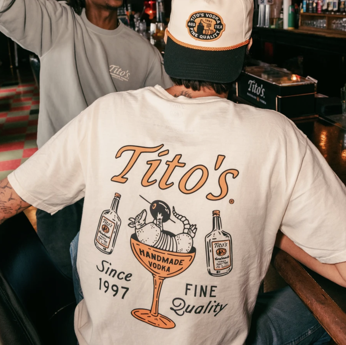 Back of a person wearing a Tito's Handmade Vodka T-shirt with illustrations of vodka bottles, a fruit bowl labeled 'Handmade Vodka,' and the text 'Since 1997 FINE Quality.' They are sitting at a bar with another person in a gray Tito's shirt and Tito's merchandise in the background.