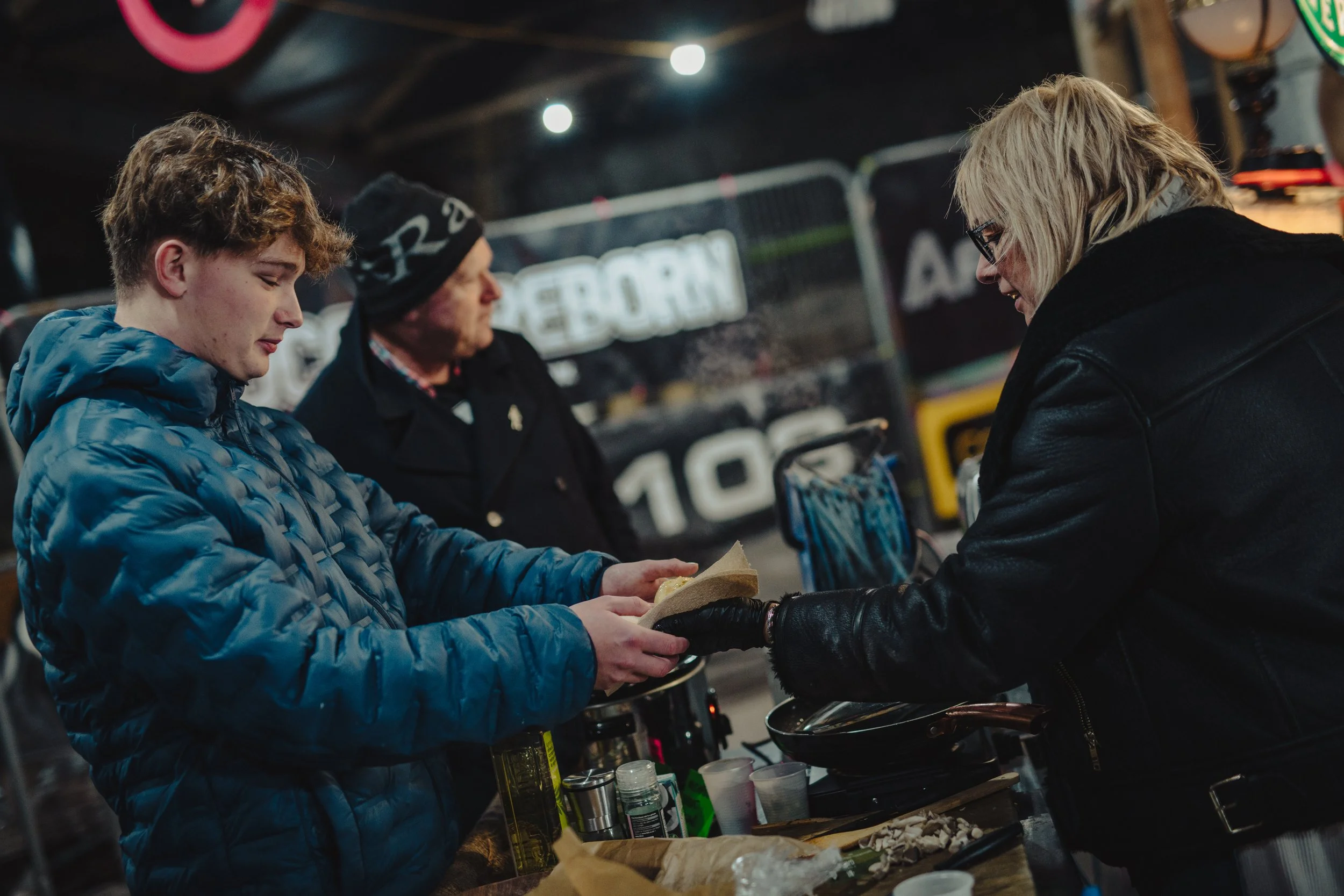 A young man in a blue jacket receives a food wrap from a woman in a black jacket at a night market or food stall. Two other people are in the background, one wearing a black beanie and the other with blonde hair, both observing the transaction. The setting appears lively with signs and equipment around.