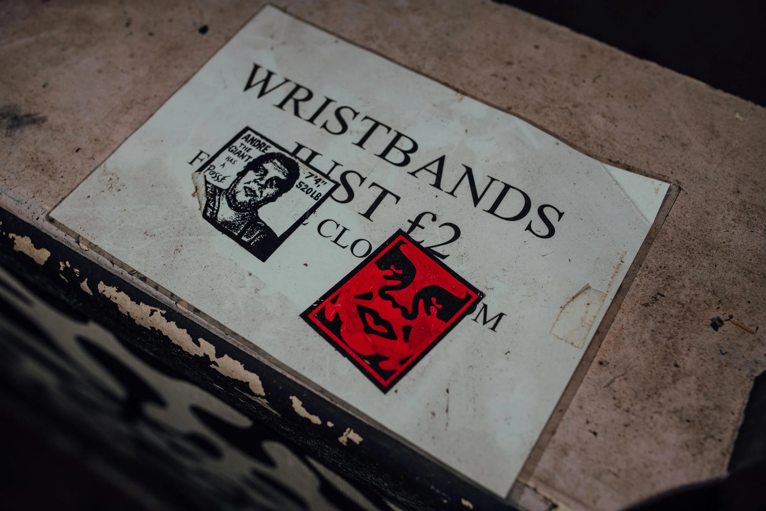 Sign on a table advertising wristbands for sale, with a black and white portrait of a man and red sticker with a black dragon illustration.