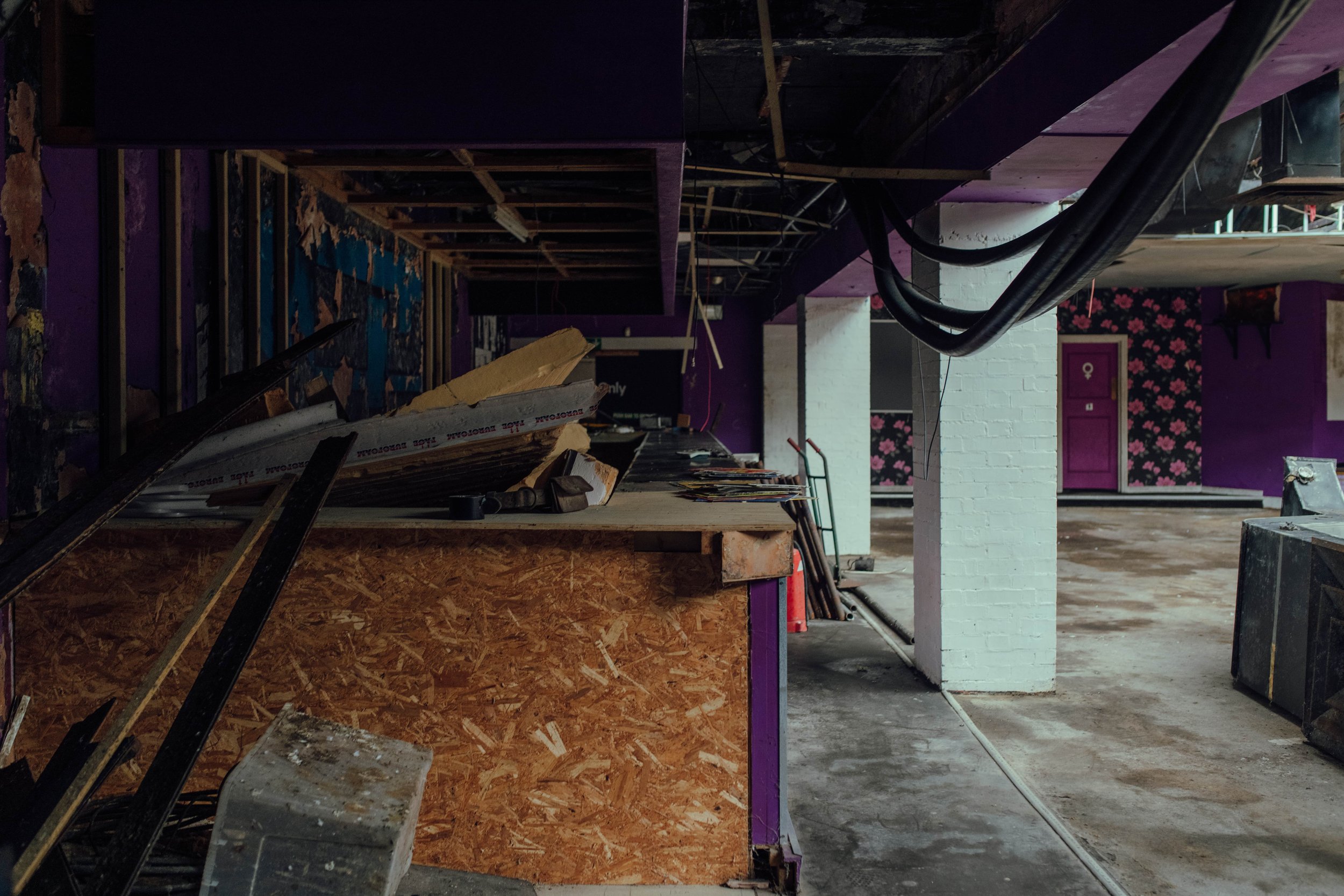 Interior of a building under renovation with construction materials, exposed ceiling, and purple walls with floral wallpaper.