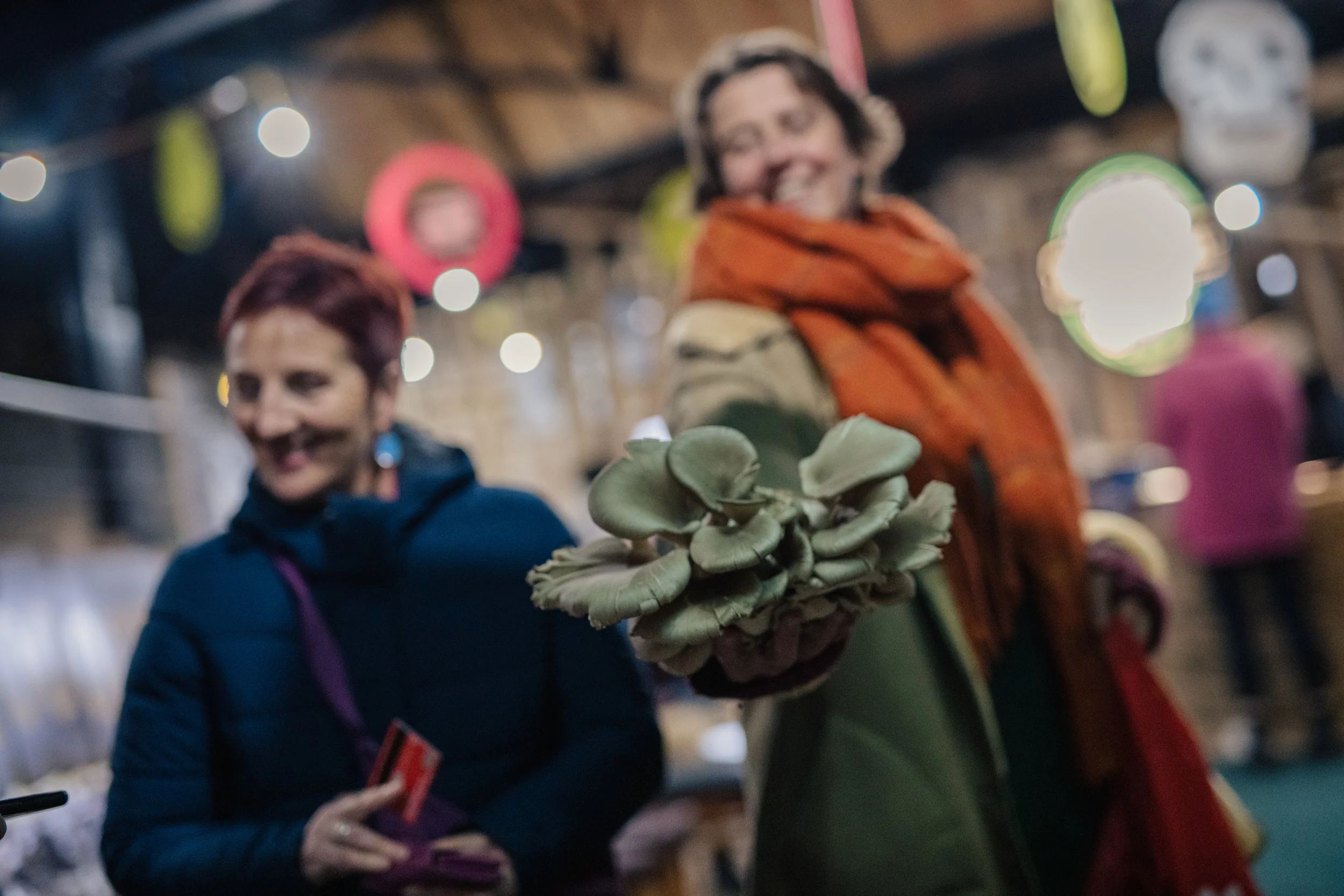 A person holding a cluster of green mushrooms, with two smiling women in the background in a festive, decorated indoor setting.