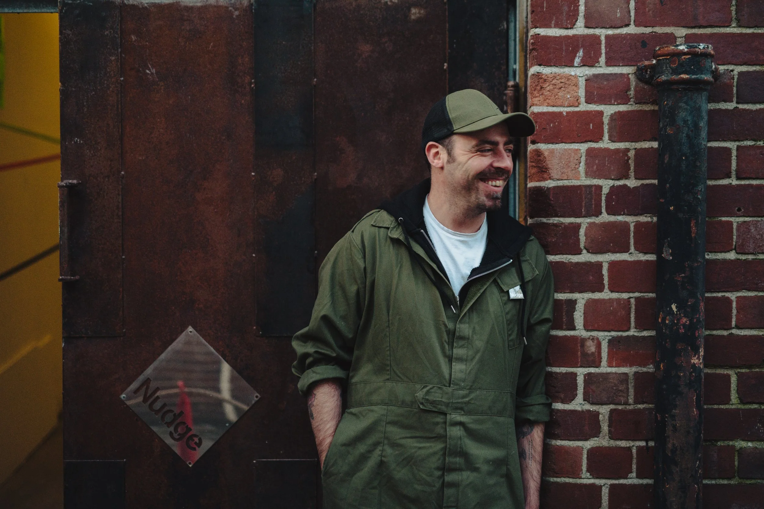 A man in a green jacket and cap smiling, standing next to a brick wall and a metal door with a 'Nudge' sign.