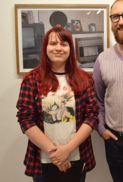 A young woman with red hair, wearing a graphic t-shirt and a red and black plaid shirt, standing indoors next to a man with glasses and a purple dress shirt. They are in front of a wall with framed pictures and records.