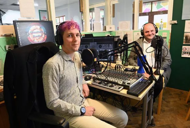 Two men in a recording studio with mixing equipment, headphones, and microphones, sitting at a table with live streaming or podcasting setup.