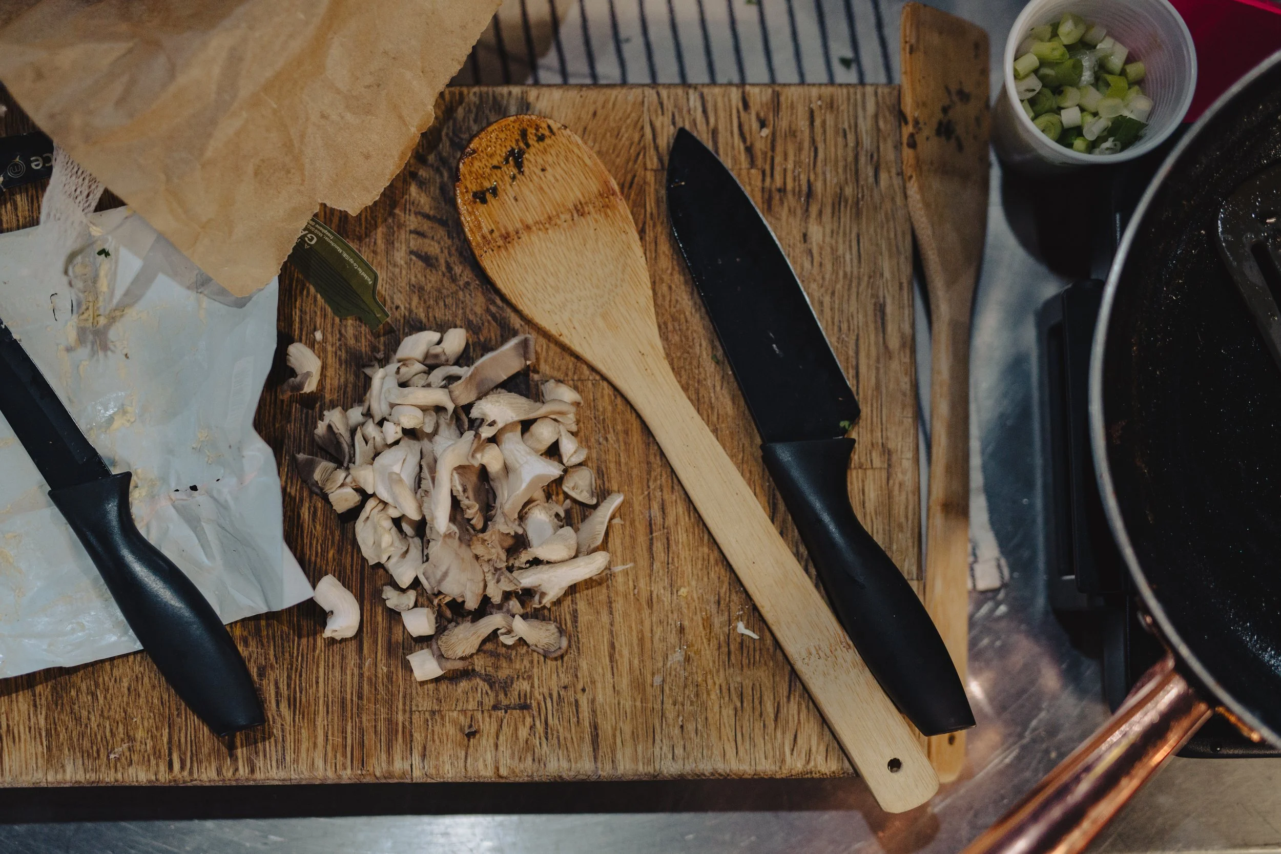 Chopped mushrooms on a wooden cutting board with a wooden spatula and a chef's knife, green onions in a small cup, and part of a black pan on a kitchen countertop.