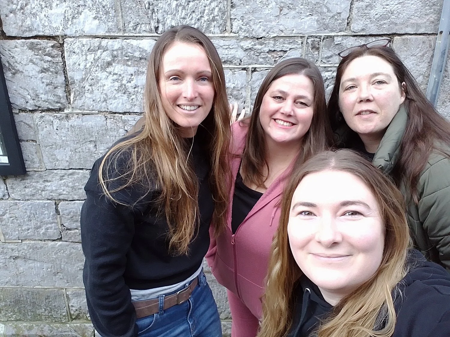 Four women taking a selfie in front of a stone wall, smiling at the camera.