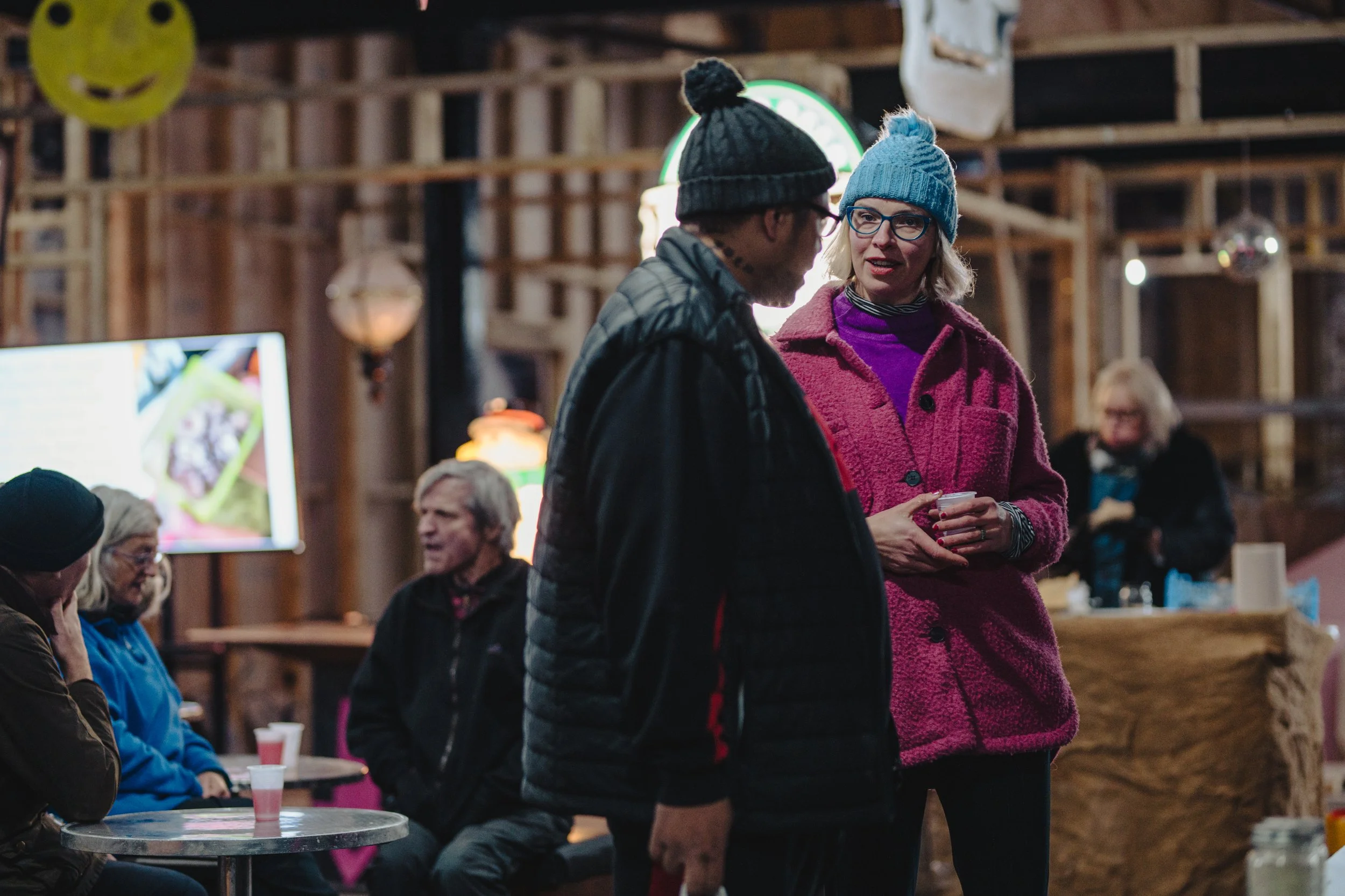Two women with winter hats and jackets talking to each other in a rustic indoor setting with other people sitting at tables in the background.