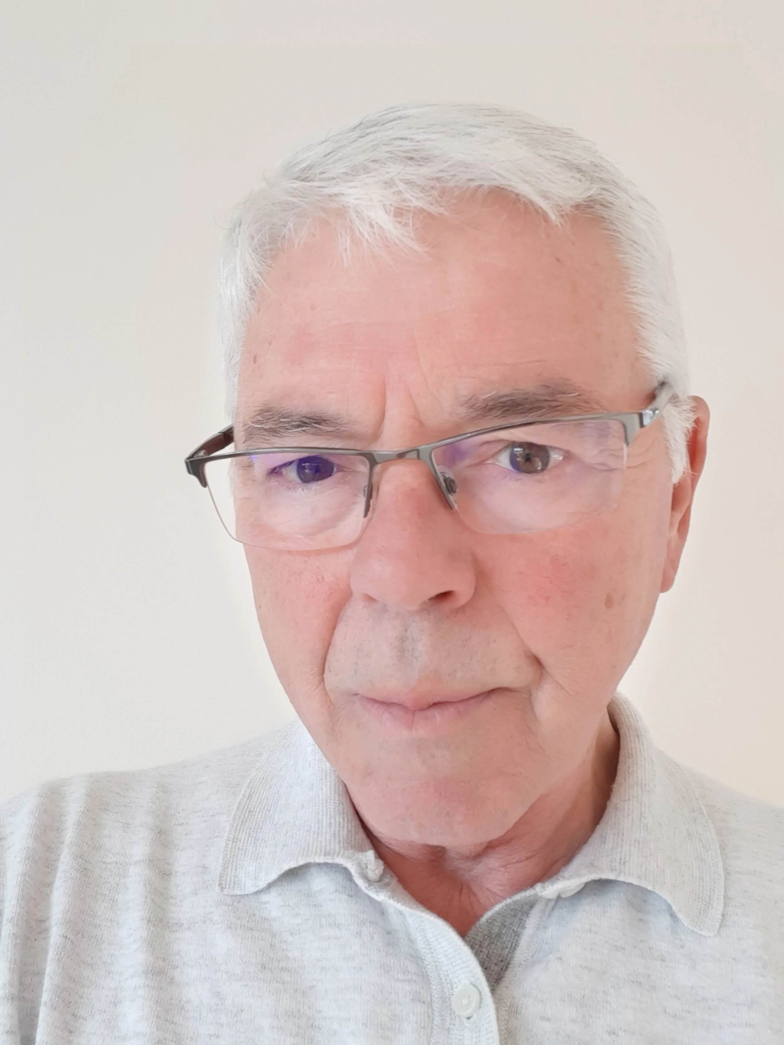 Close-up portrait of an older man with gray hair and glasses, wearing a light gray collared shirt, looking directly at the camera against a plain light background.
