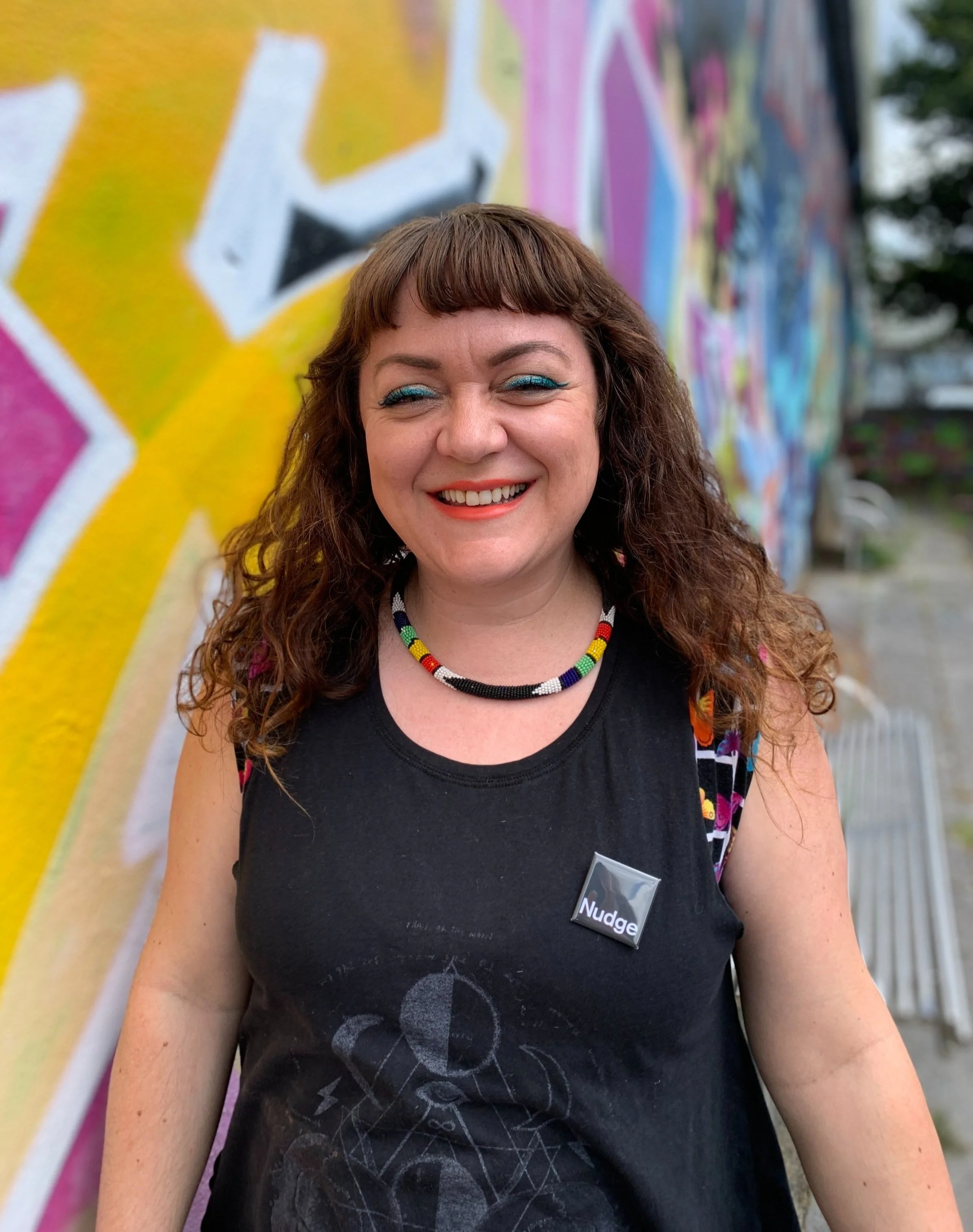 A woman with curly brown hair, wearing bright blue eyeshadow, a black sleeveless shirt with a design, a multicolored beaded necklace, and a name tag that says 'Nudge' stands smiling in front of a colorful graffiti wall.