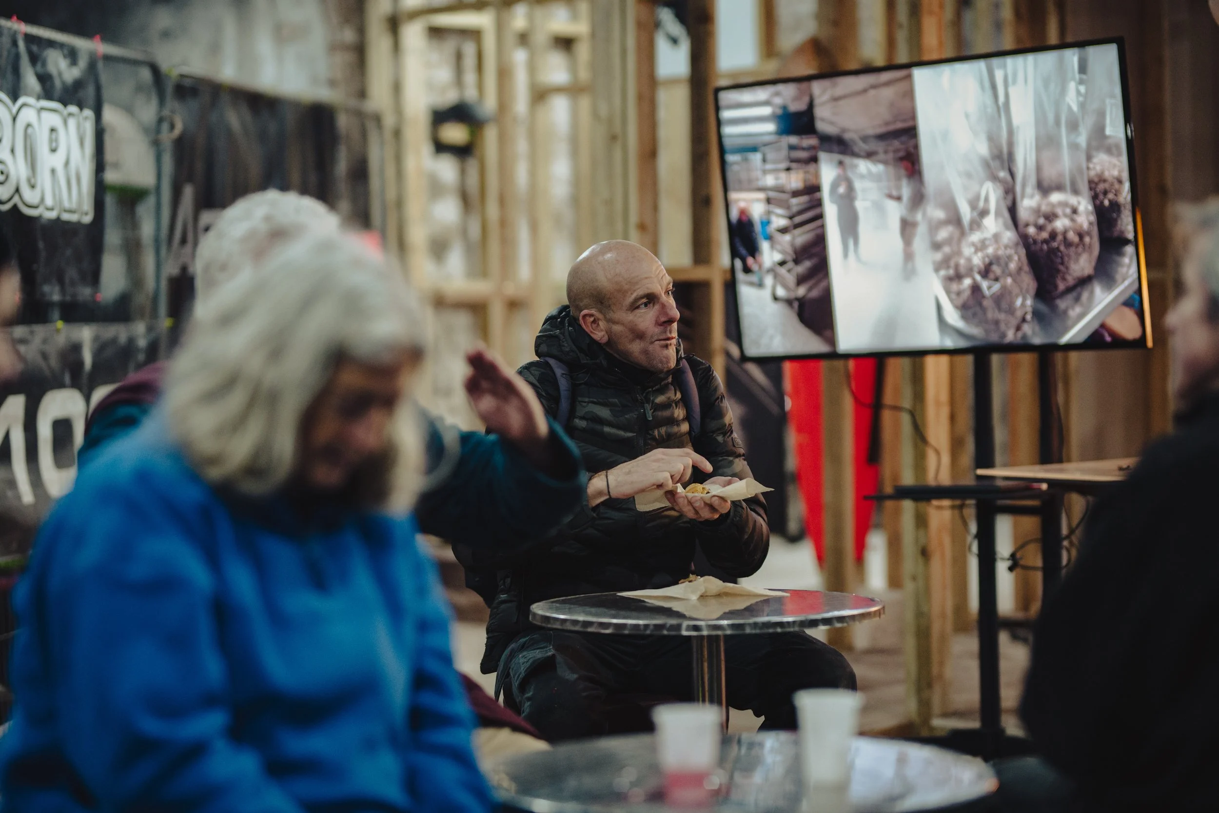 Group of people sitting at tables in a rustic indoor setting, with a man in the middle eating and a large TV screen behind him displaying images of food and people.