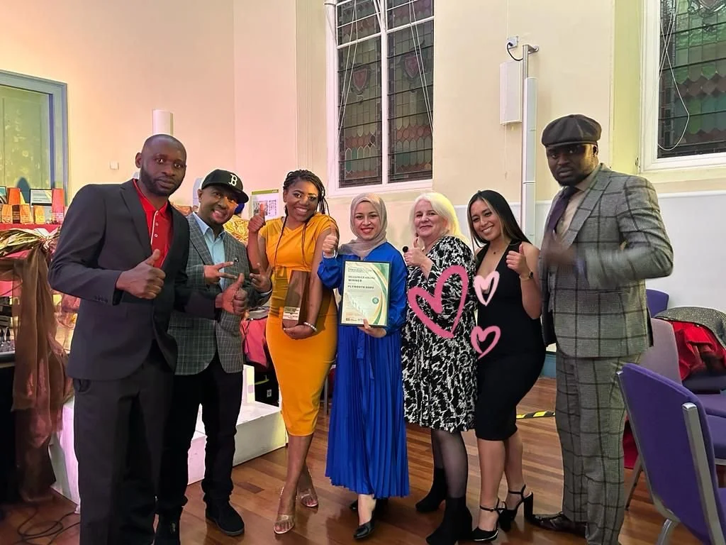 Group of seven diverse people standing indoors in a celebratory setting, holding a certificate and posing for a photo.