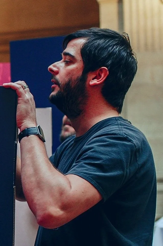 A man with dark hair and a beard, wearing a dark t-shirt, looks at a vertical surface, possibly a screen or display, in an indoor setting.