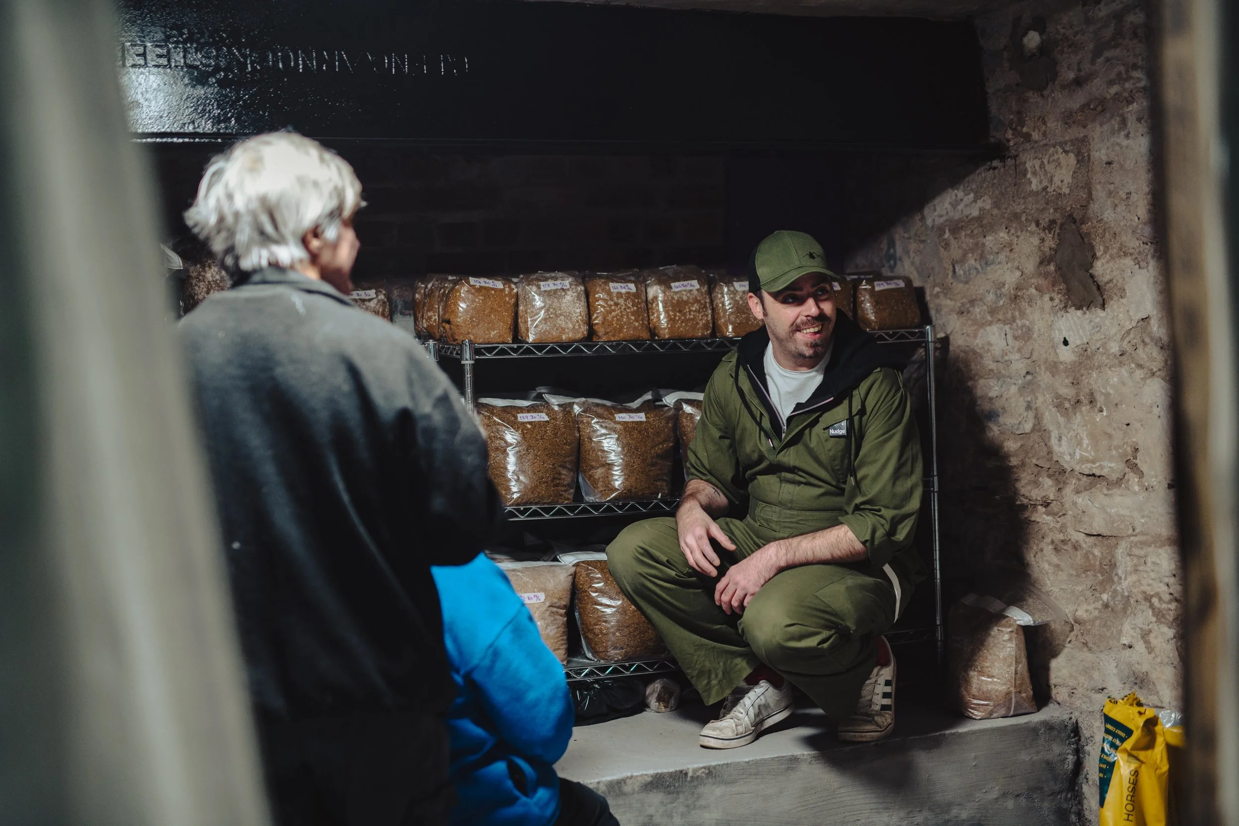 A man in a green cap and jacket crouching and smiling while talking to an older person with white hair in a cellar with shelves of packaged dried herbs or spices.
