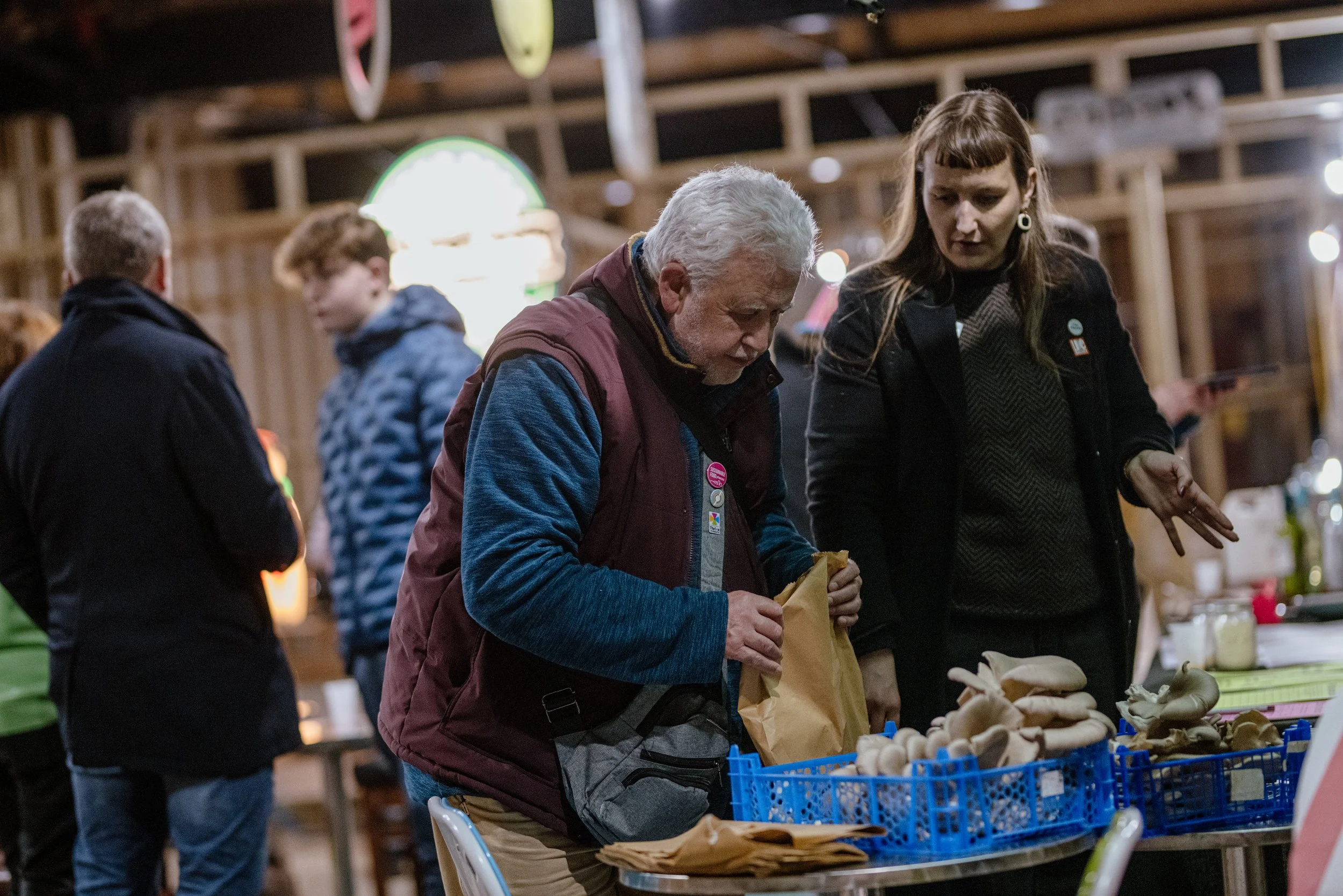 A man and a woman standing at a table with mushrooms in blue crates, in an indoor market setting.