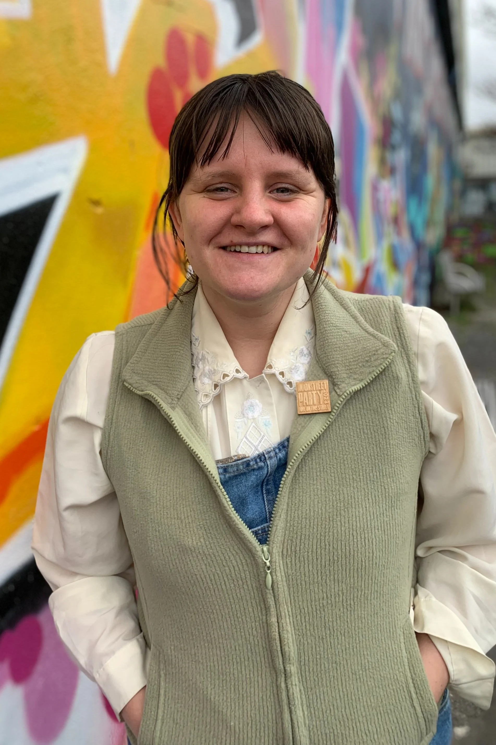 Young woman with short brown hair, smiling, standing in front of a colorful graffiti wall in an outdoor setting.