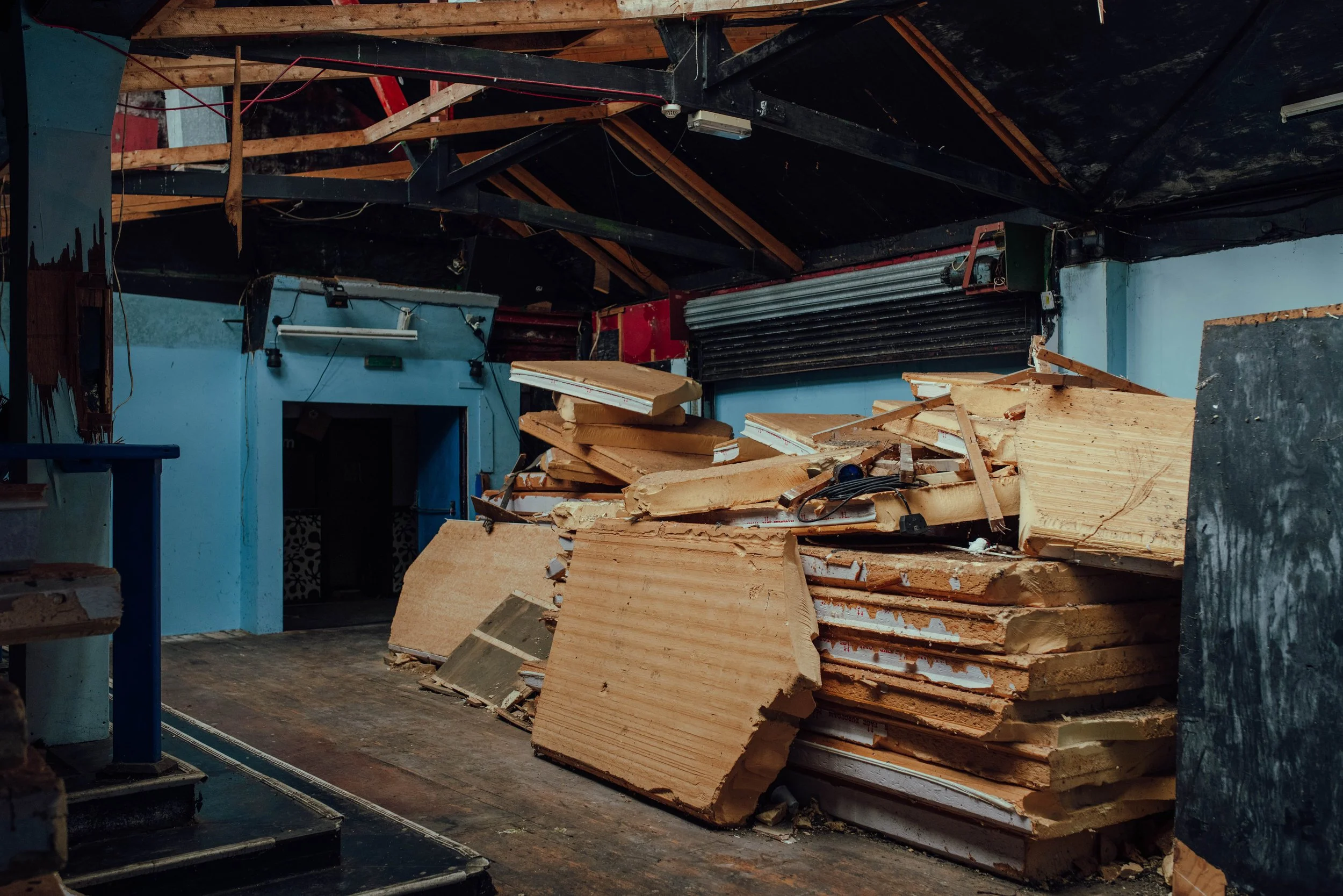 Interior of a woodworking shop with a pile of stacked wooden boards, some damaged, and tools scattered around. The ceiling is partially exposed with wooden beams and a black wall or ceiling covering.