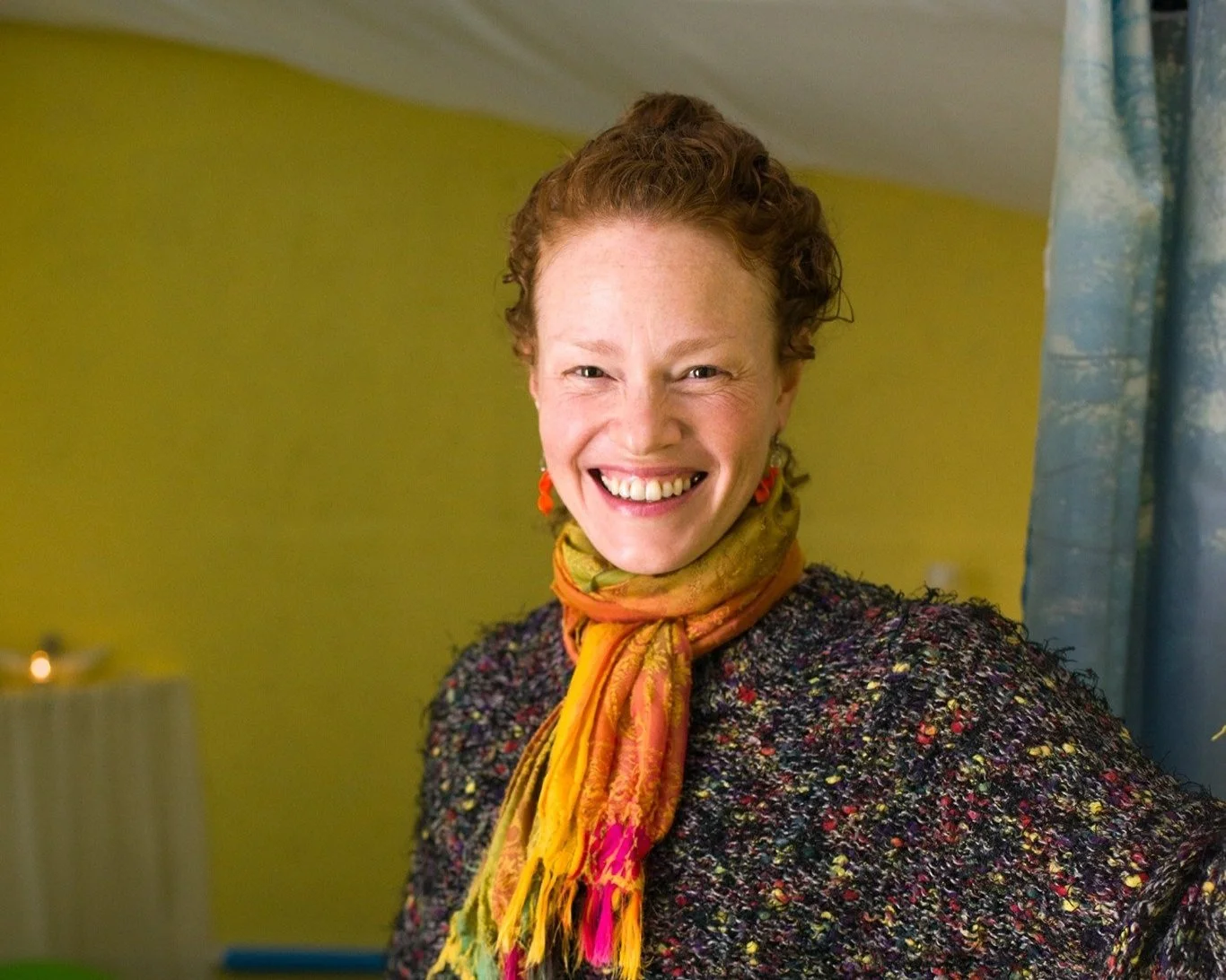 A woman with short curly red hair and earrings smiling in a room with a yellow wall and blue curtains.