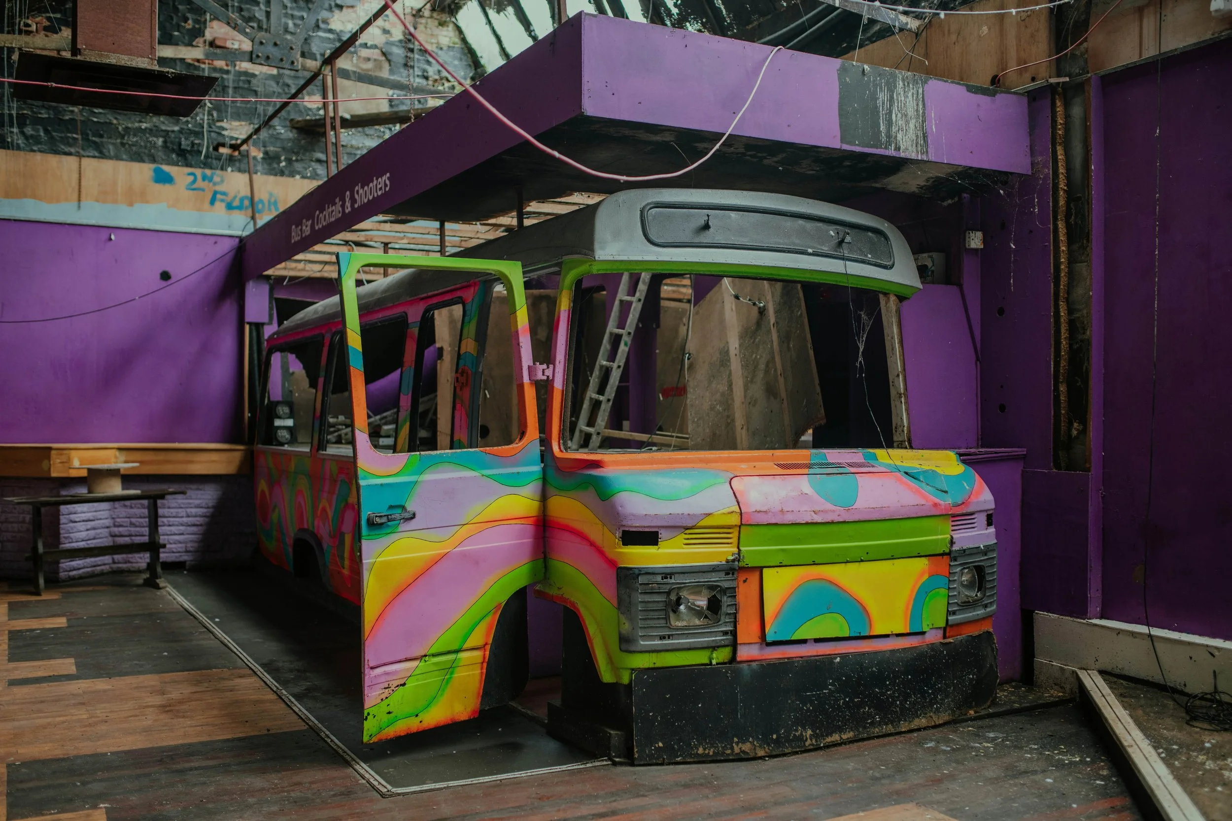 Colorful, psychedelic-painted bus front and side panels inside an abandoned, dilapidated building with purple walls.