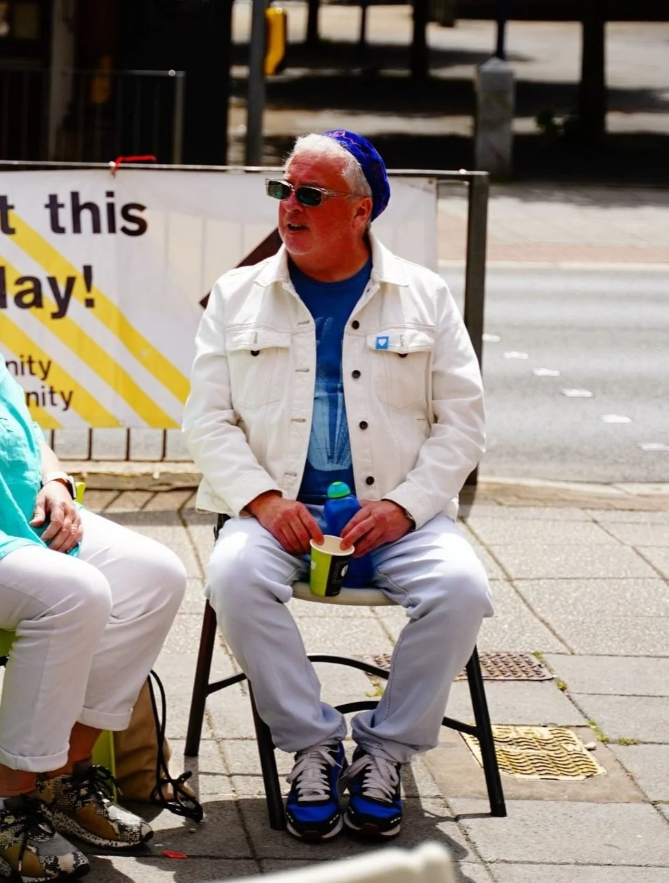 A man sitting outdoors on a white chair, wearing sunglasses, a blue hat, a white jacket, and white pants. He is holding a green cup and engaged in conversation.