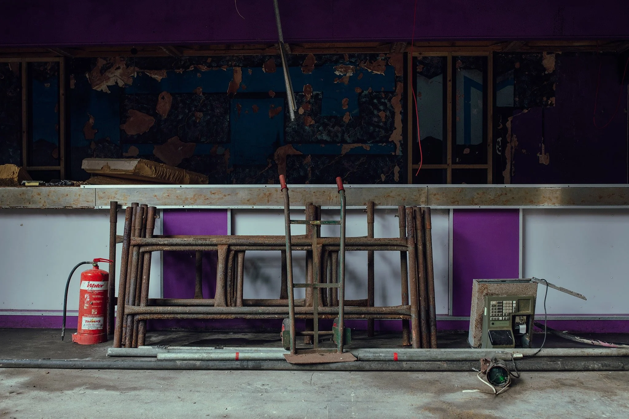 Construction site with metal scaffolding, a fire extinguisher, and an old electronic device on a concrete floor, with unfinished wall and wall framing in the background.
