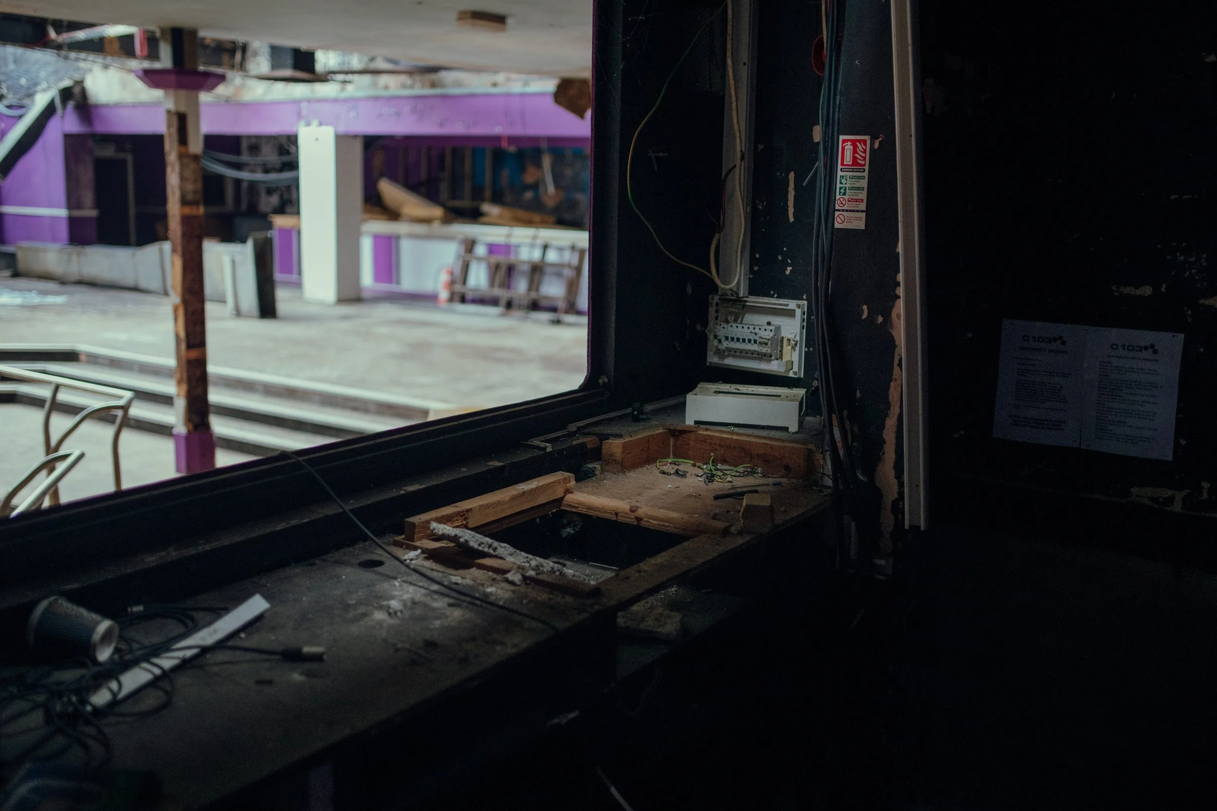 Interior of a damaged building with broken or missing window, exposed wiring, and debris on the surface, overlooking a partially demolished or under renovation space with purple and white walls.