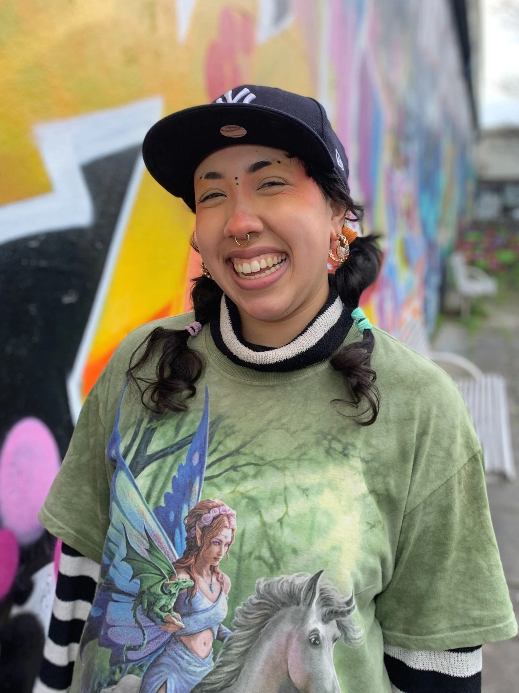 Young woman with a big smile, wearing a black New York Yankees cap, a graphic T-shirt with a fairy and horse, and standing in front of a colorful graffiti wall.