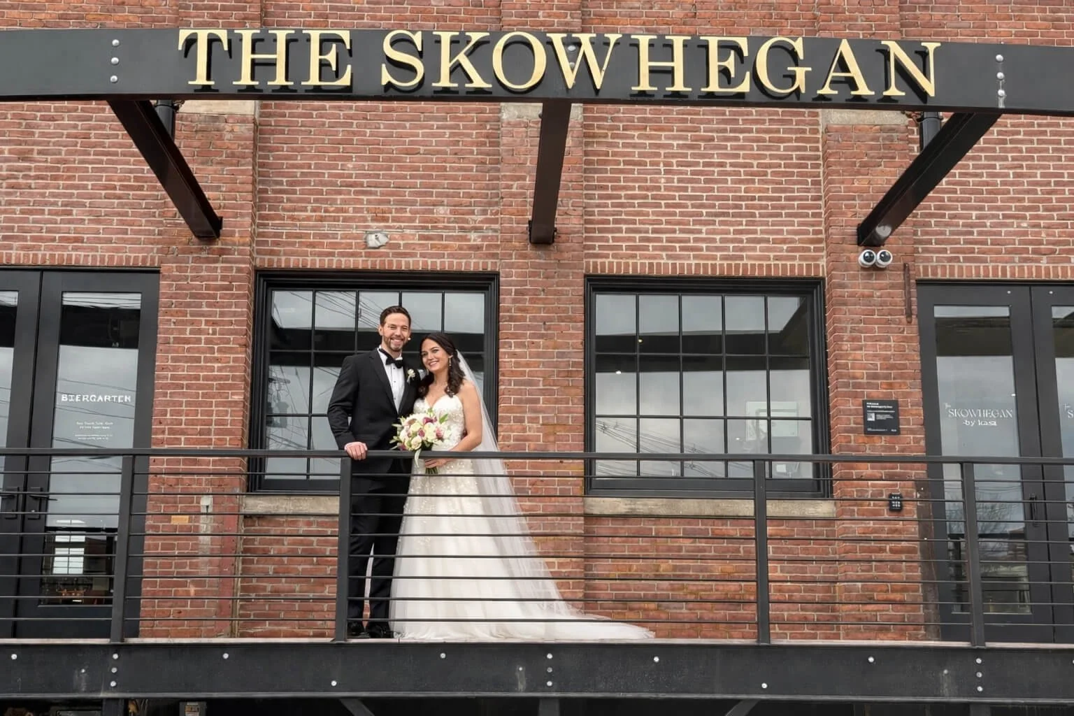 A newlywed couple in wedding attire standing on a balcony in front of a brick building, with a sign reading 'THE SKOWHEGAN' above them.
