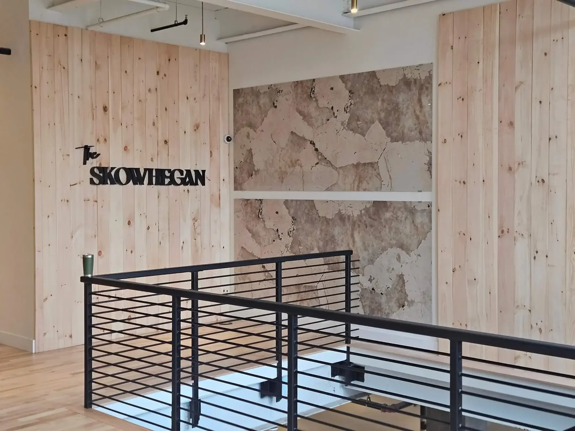 Interior of a modern building with wood-paneled walls, an exposed ceiling with track lighting, and a section of cracked marble wall. A black metal railing surrounds a staircase opening in the floor.