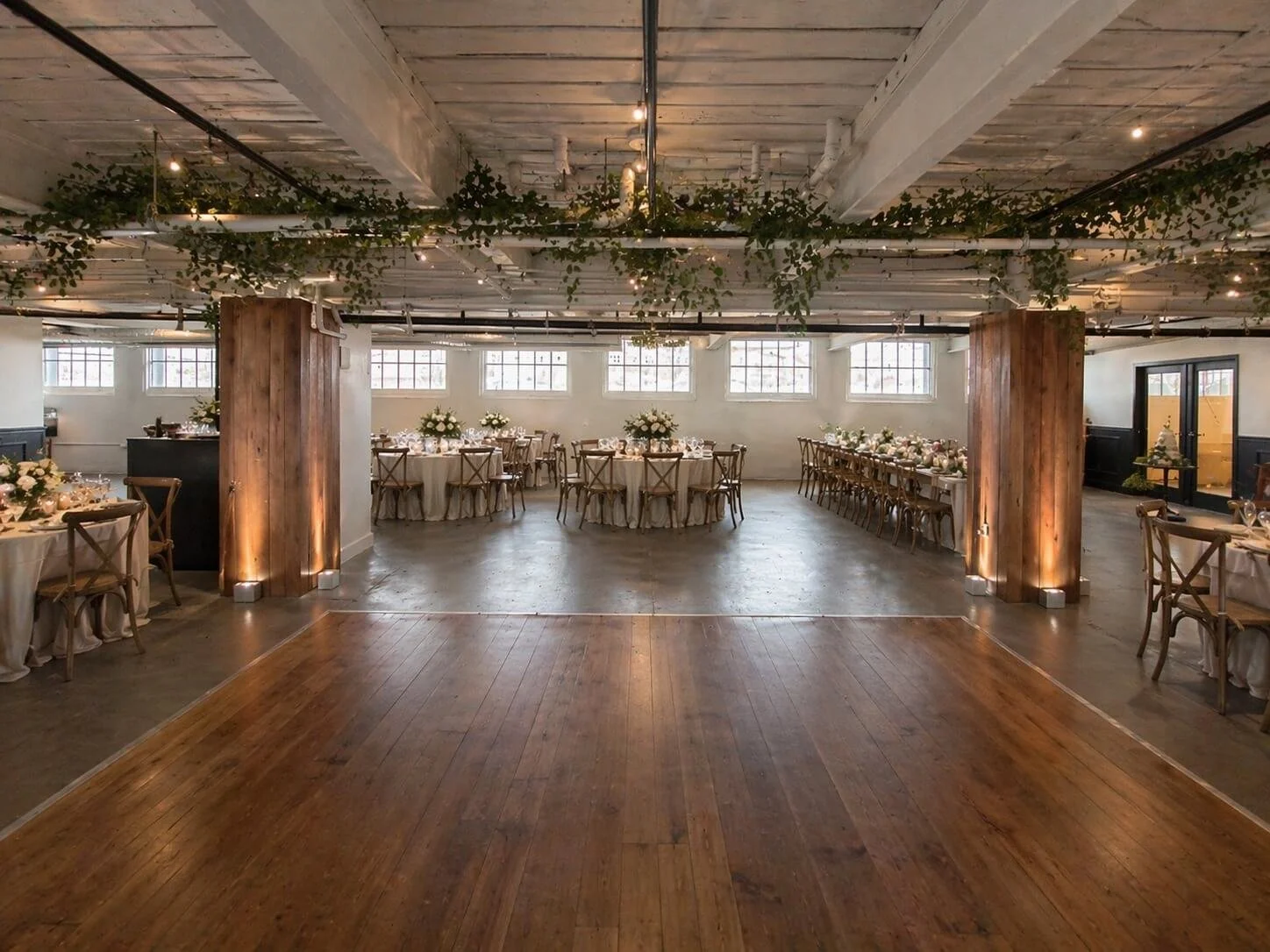 Wedding reception hall decorated with round tables and long banquet tables adorned with white flowers, green foliage hanging from the ceiling, and warm lighting. The flooring combines wood and concrete with large industrial windows in the background.