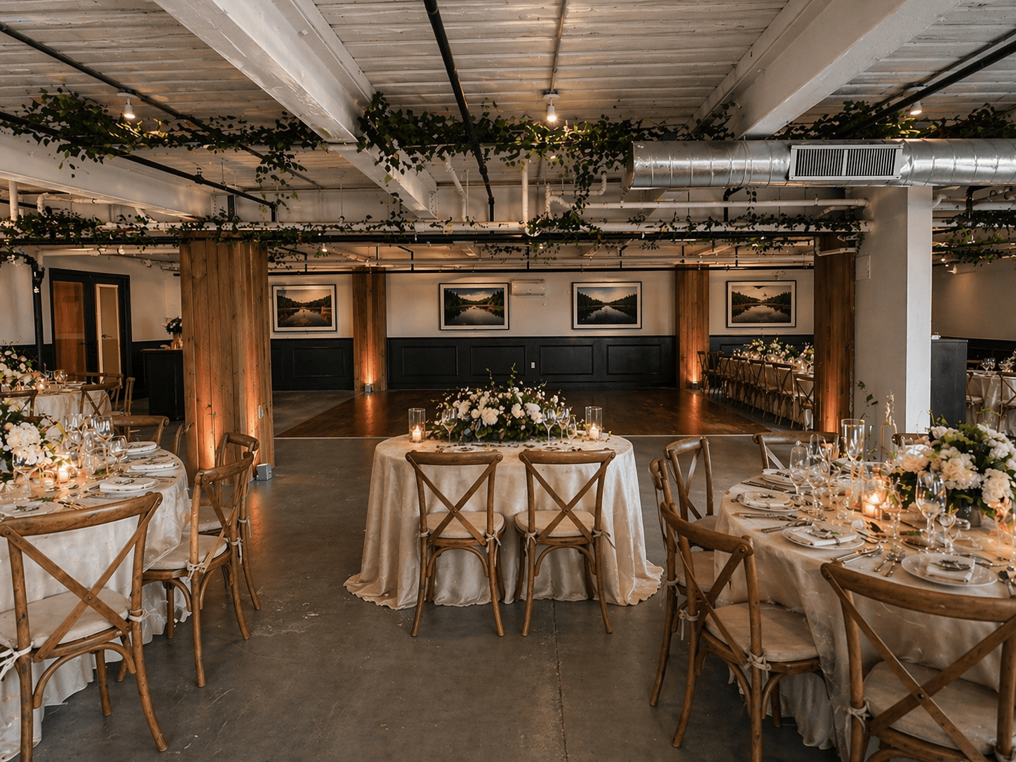Elegant event space decorated with round tables covered in cream tablecloths, featuring floral centerpieces with white flowers and candles, wooden cross-back chairs, and framed landscape photographs on the wall, with wooden beams and greenery decorating the ceiling.
