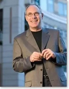 A smiling man in glasses wearing a gray suit jacket and black shirt, standing outdoors near a building with columns.