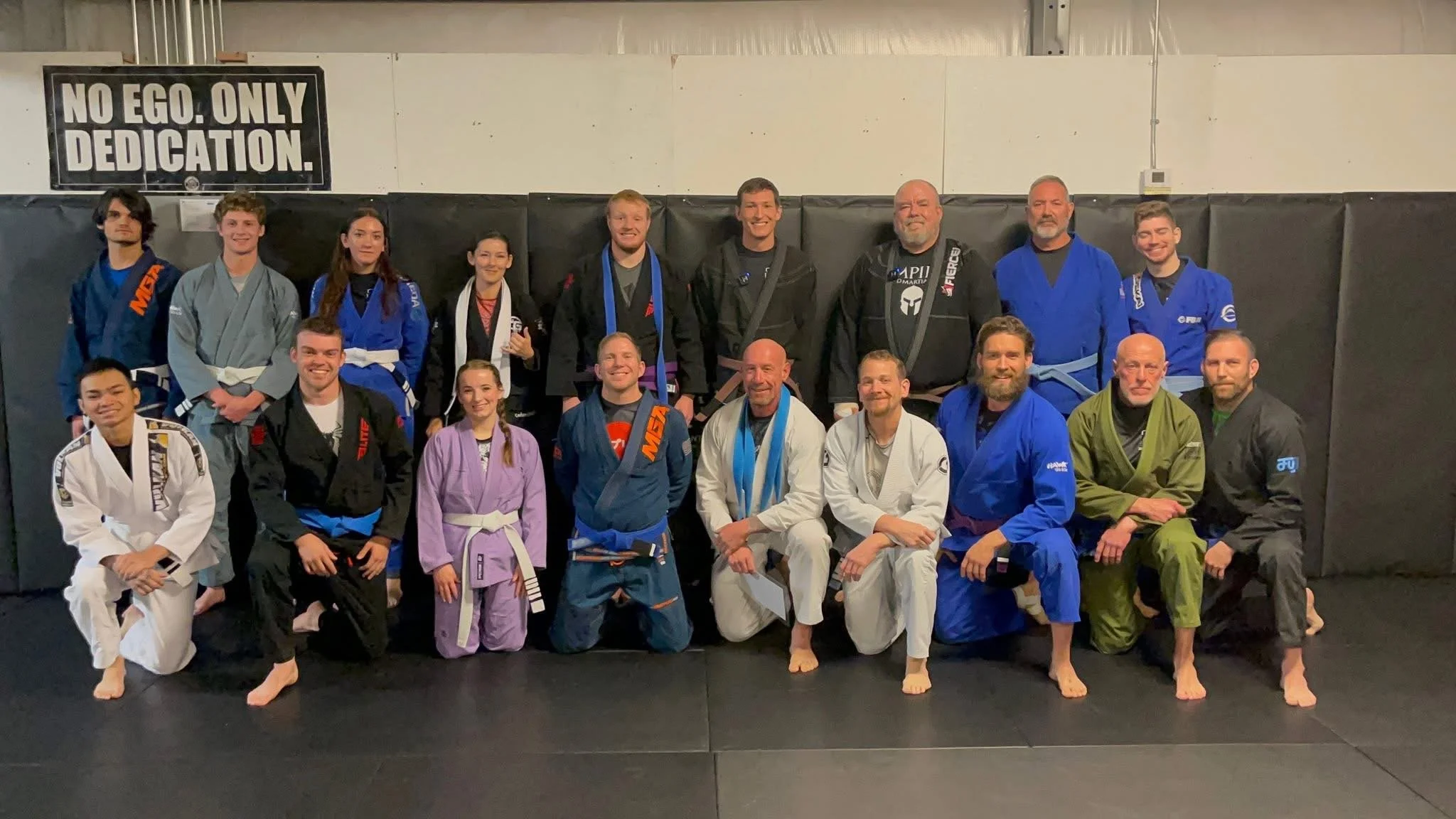 Group of people, including men and women, dressed in Brazilian Jiu-Jitsu gis and belts, posing for a photo in a martial arts gym. There is a sign on the wall that reads 'No Ego. Only Dedication.'