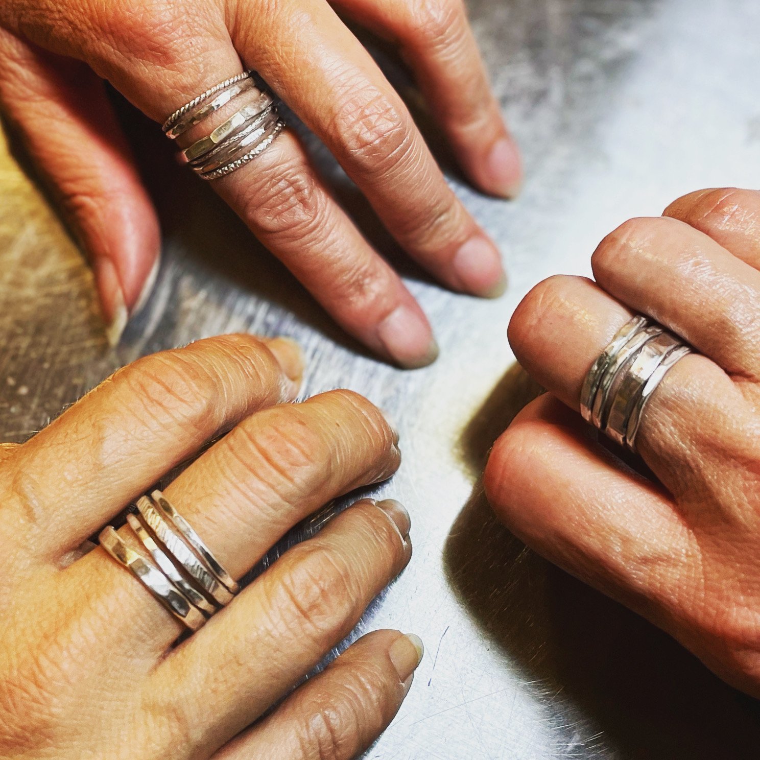 Three hands with silver rings on fingers, placed on a metallic surface.