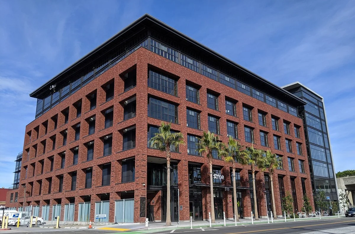 Stripe’s headquarters: a six-story red-brick office building with tall windows and palm trees along the sidewalk, under a blue sky
