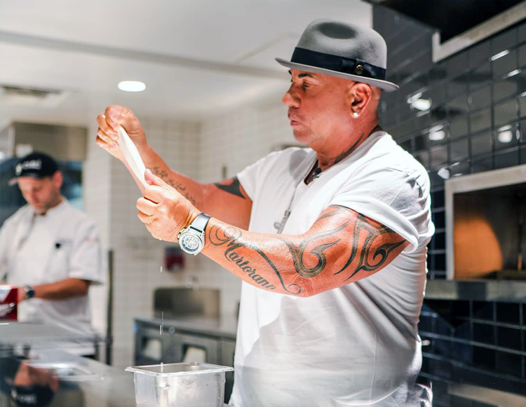 Steve Martorano in a white T-shirt and grey fedora, tossing pasta dough in his restaurant kitchen