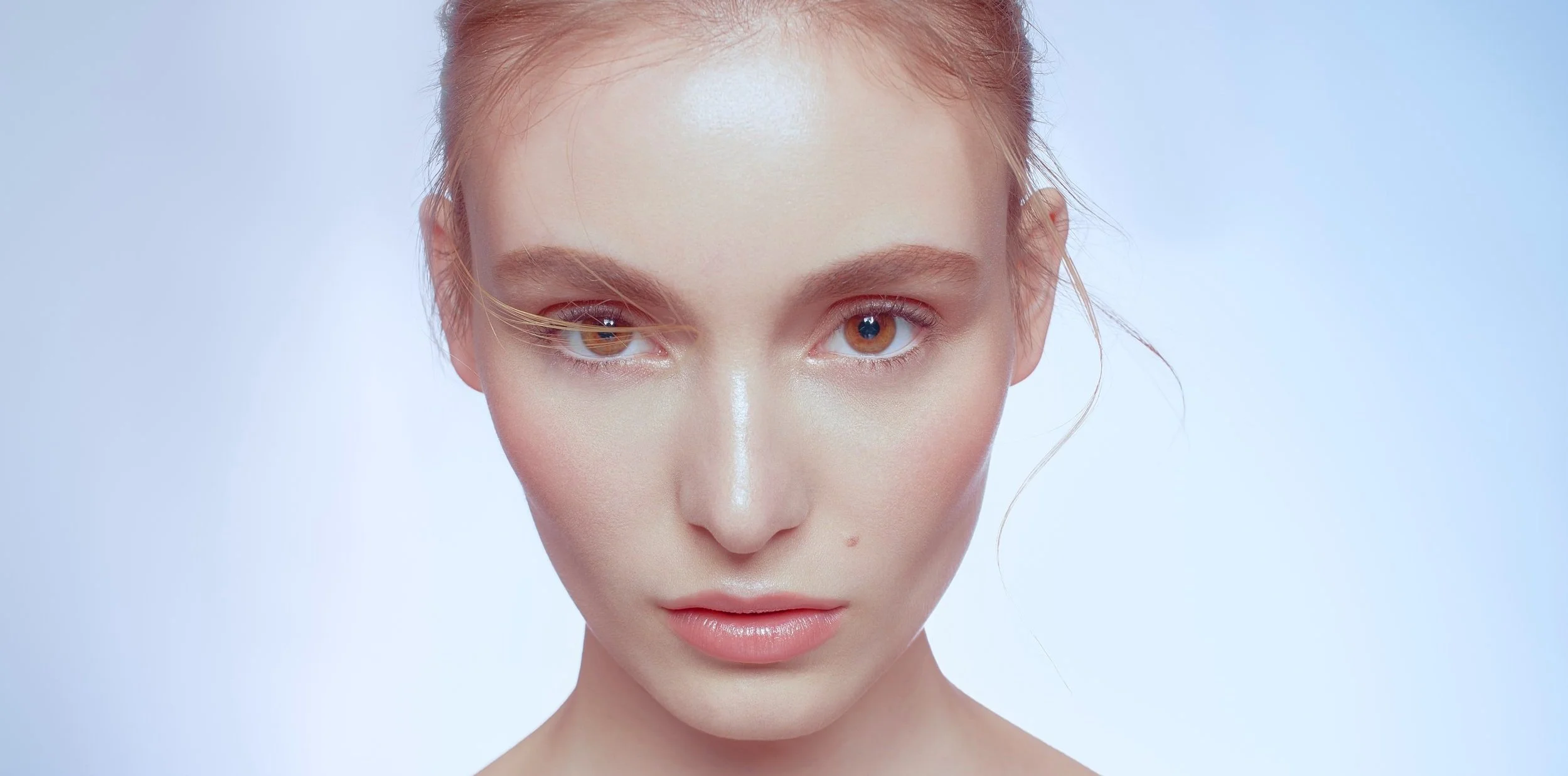 Stunning shot of a woman looking straight into the camera. Her amber eyes and dewy skin pop against the blue background. Photo by advertising photographer, Nathanael Filbert. 