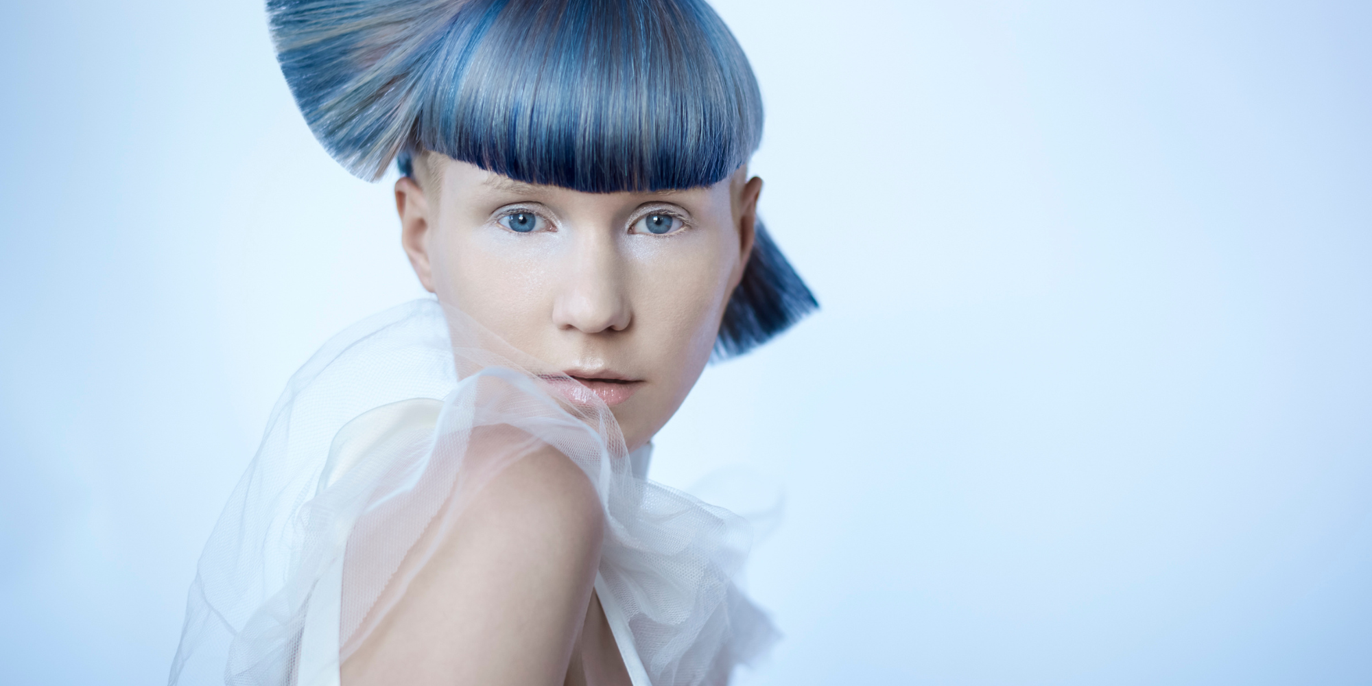 The future is here in this bright photograph of a woman with ice blue eyes and matching blue hair is set against a light blue backdrop, the texture of her wardrobe adding to the energy. Shot by Chicago based professional photographer, Nathanael Filbe