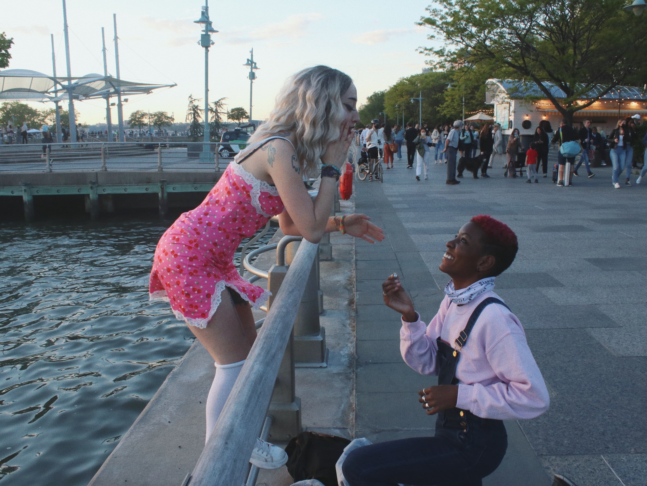 Two people near a waterfront, one kneeling and holding a ring, proposing to the other, who is covering their mouth in surprise. Background shows a promenade with people walking, trees, and waterfront structures.