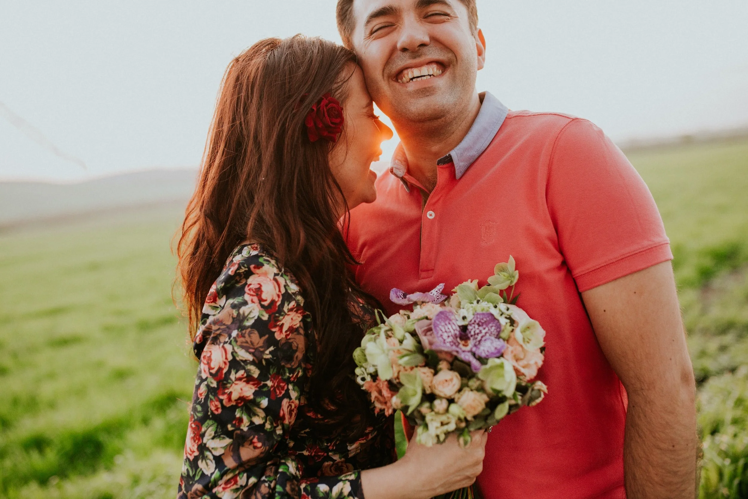 Smiling couple in a field holding a bouquet of flowers, woman wearing a floral dress and man in a red shirt.