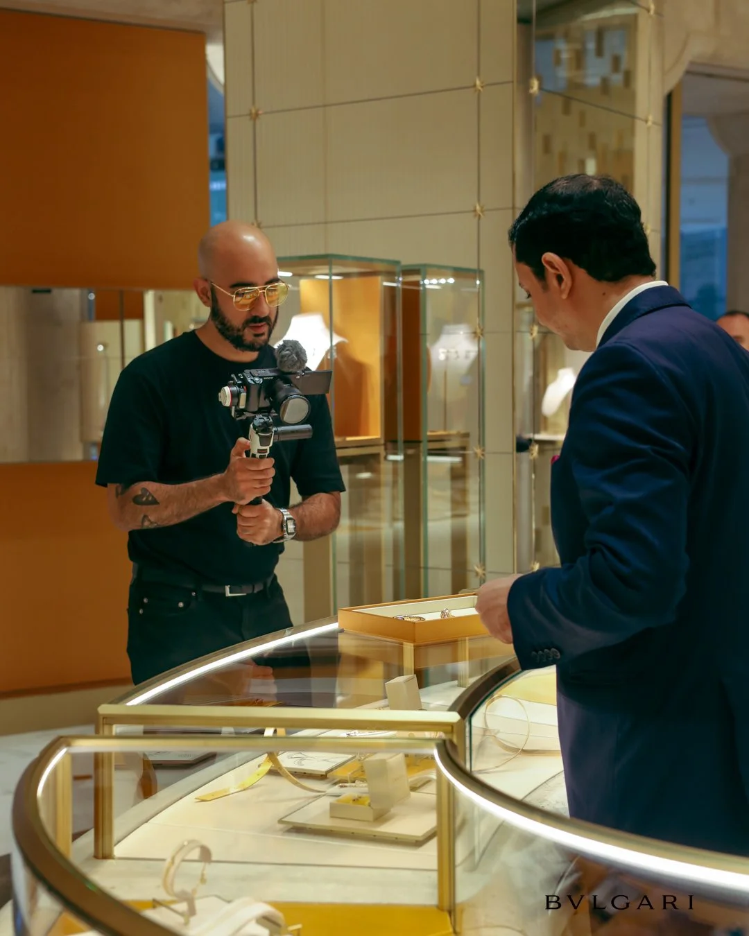 Dos hombres, uno con barba, gafas de sol y camiseta negra, está filmando con una cámara en una joyería, mientras el otro, vestido con traje azul, mira unas joyas en una vitrina de exhibición.