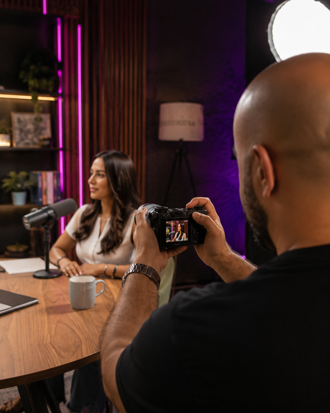 Un hombre toma una fotografía de una mujer durante un Podcast en un estudio con iluminación morada y decoraciones de estanterías.