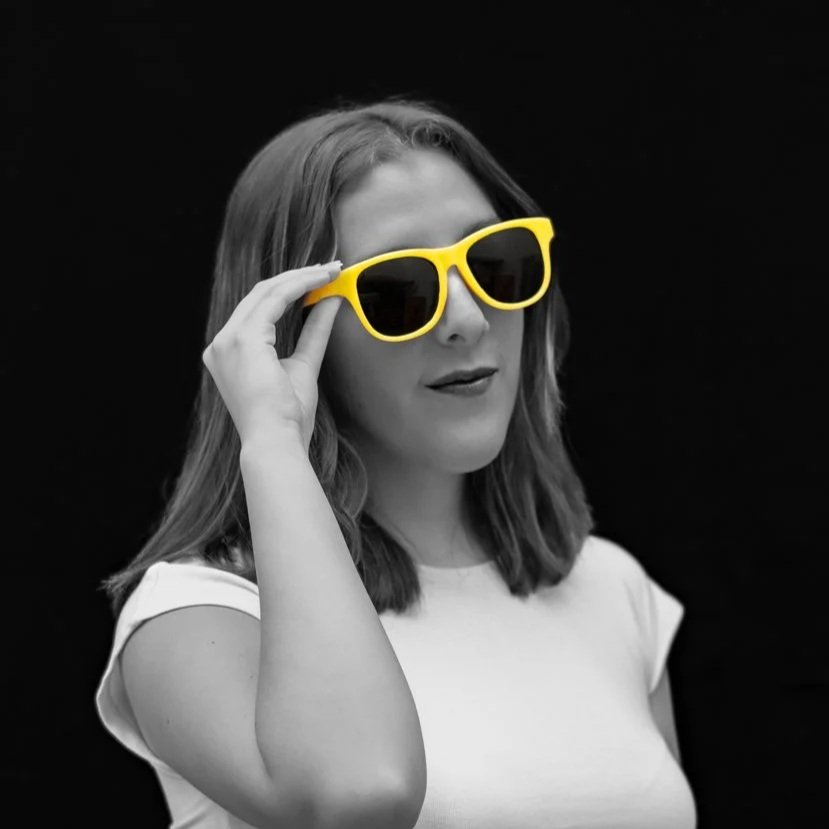 Mujer con gafas de sol amarillas, en fondo negro, en tonalidad en escala de grises, sonriendo ligeramente.