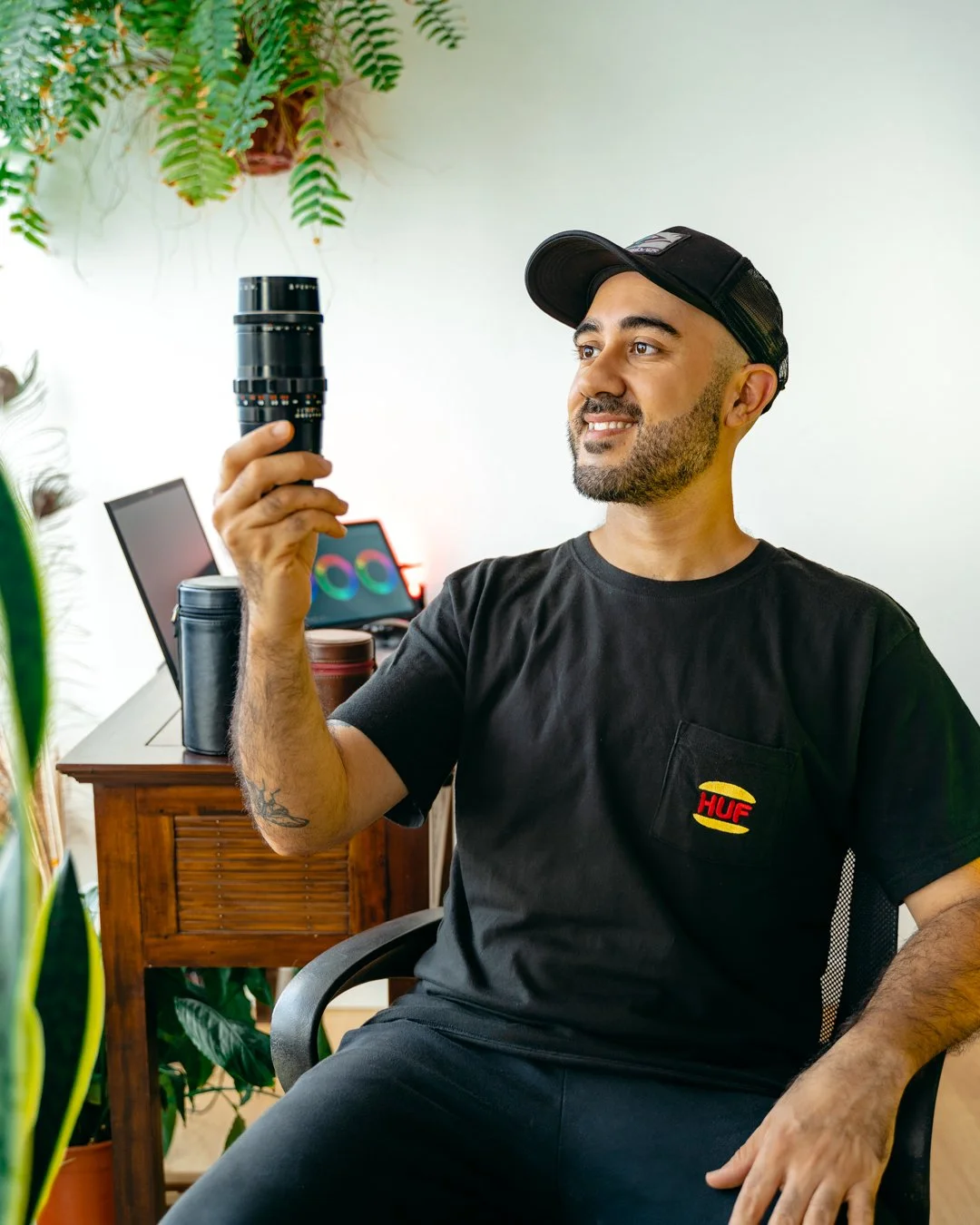 Hombre sentado en una habitación rodeada de plantas, sosteniendo un lente fotográfico. Tiene barba, lleva gorra y camiseta negra con logotipo de HUF, y está sonriendo.