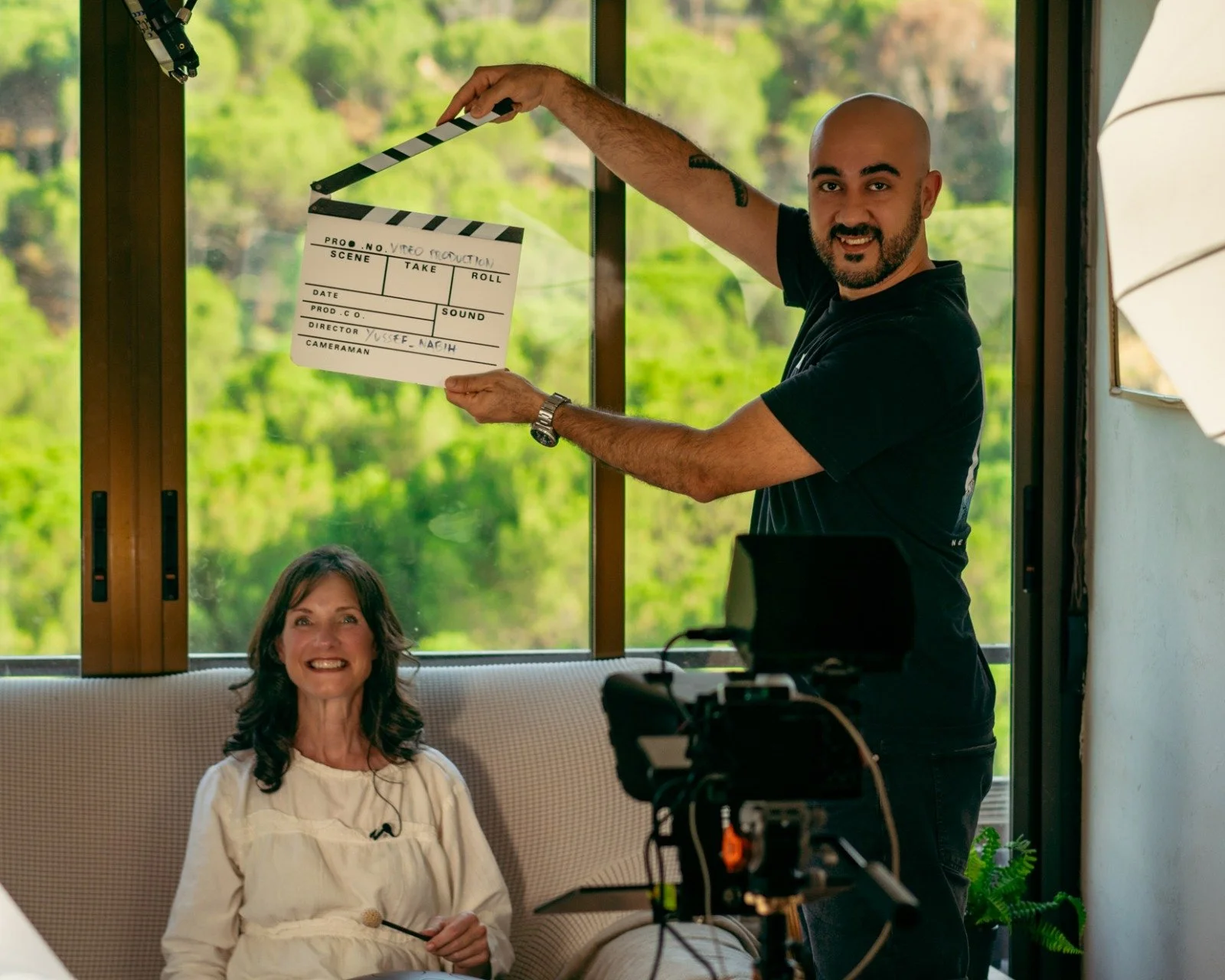 Persona sosteniendo un cartel de producción de video en un entorno interior con ventanas y naturaleza visible afuera, mientras otra persona está sentada en un sofá sonriendo.