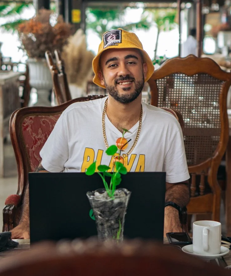 Hombre sonriendo sentado en restaurante, lleva gorra amarilla, camiseta blanca y cadenas doradas, hay una planta en la mesa y una taza con café.